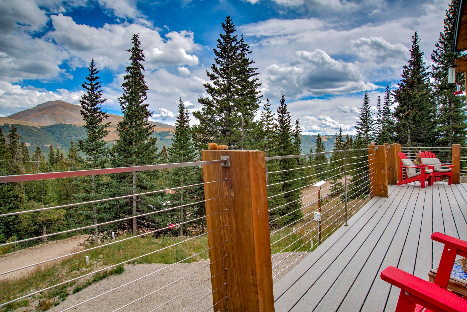 Wooden deck overlooking a mountain landscape, featuring cable railings, pine trees, and red chairs.