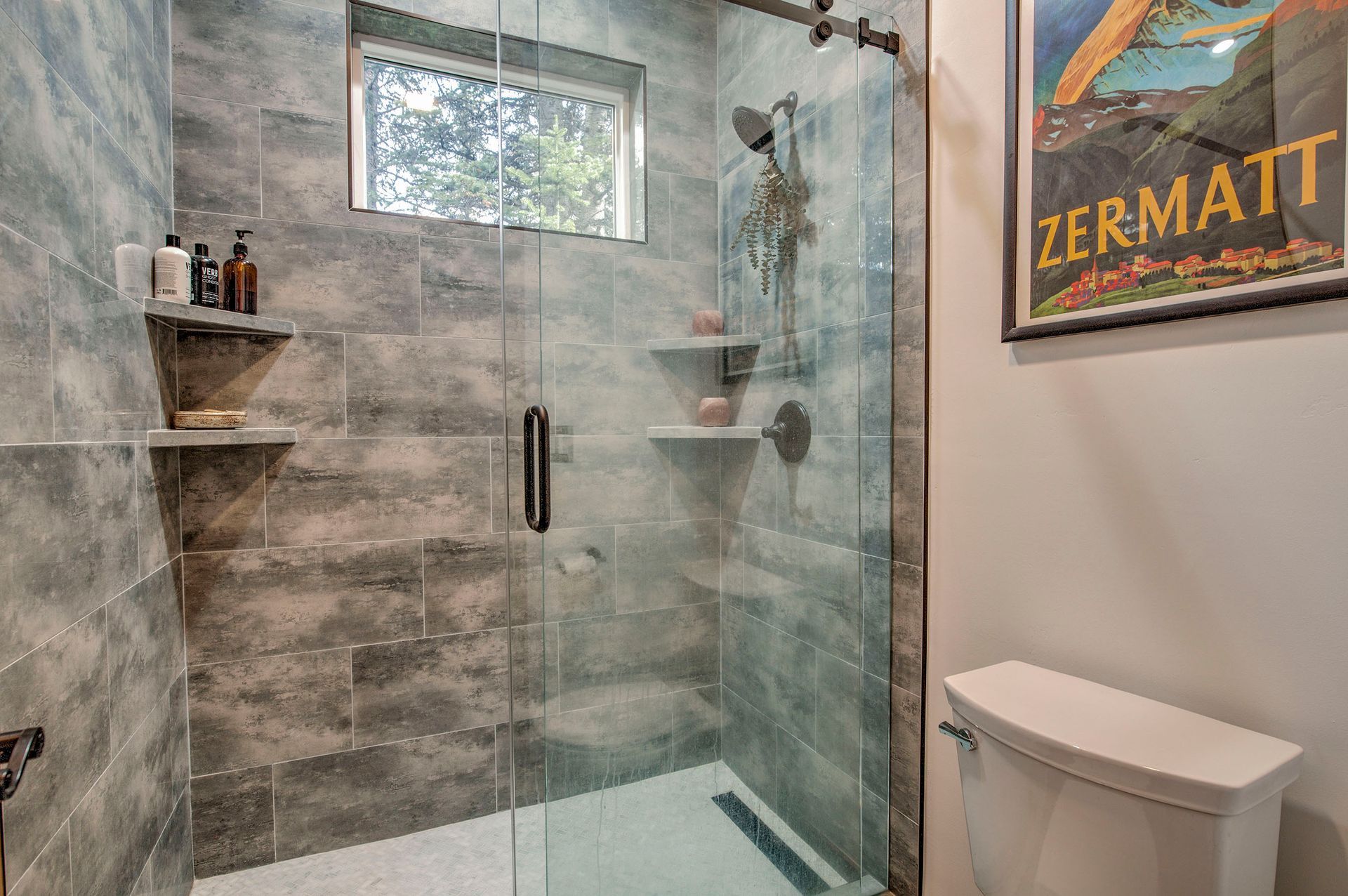 Modern bathroom with glass shower, gray tile walls, shelves holding toiletries, and a toilet.