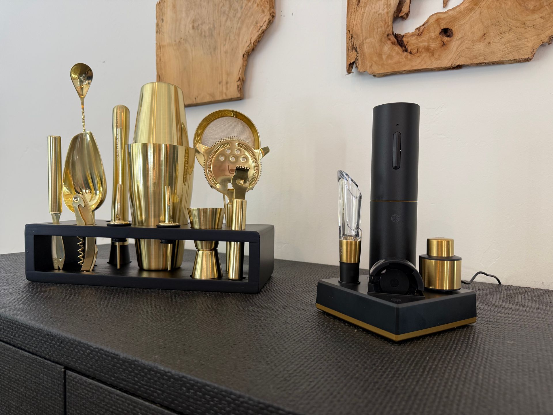 Two bar sets with gold and black accessories on a dark surface, near wood art.