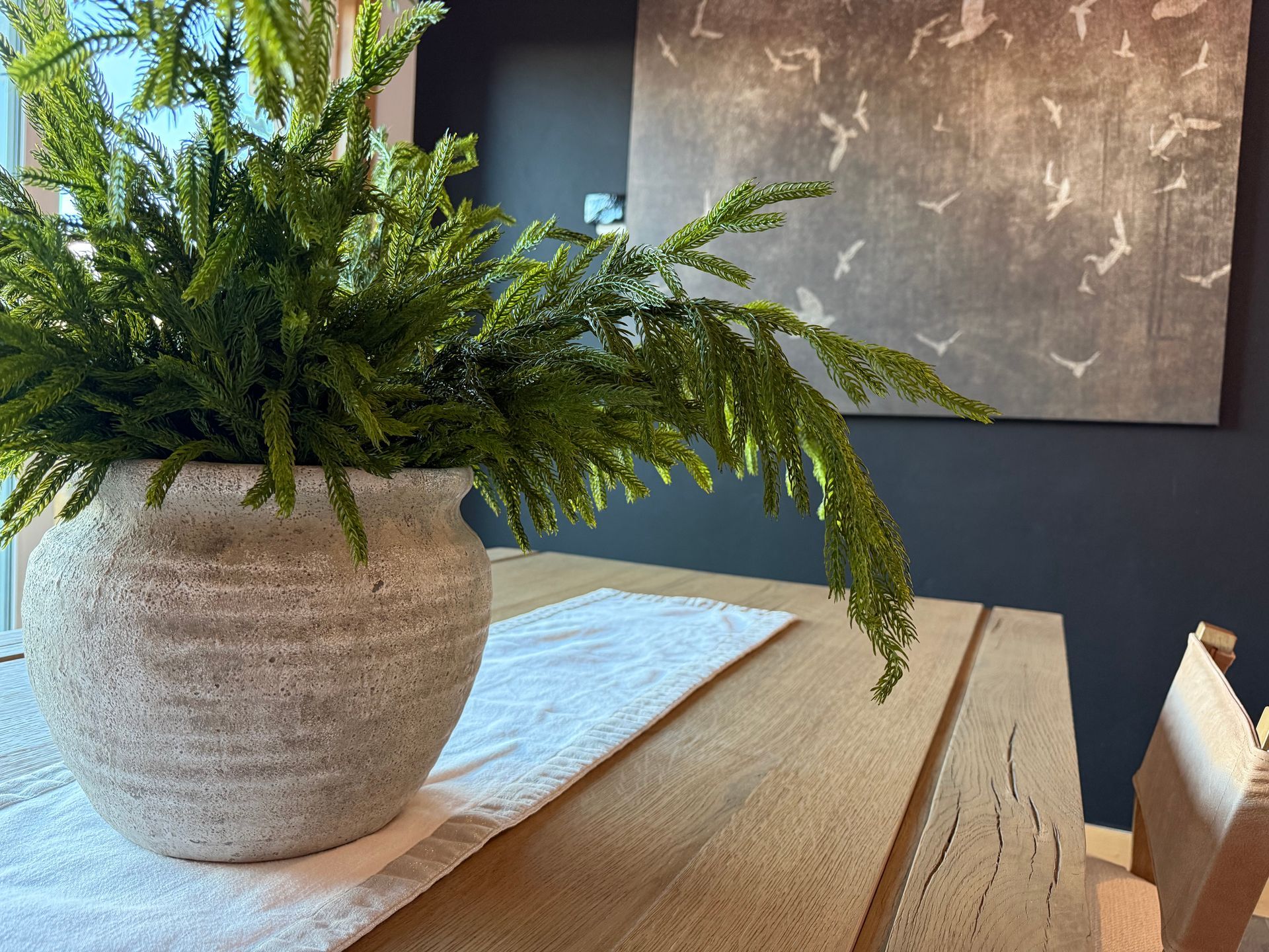 Green fern branches in a light stone pot on a wooden table with a white runner and wall art.