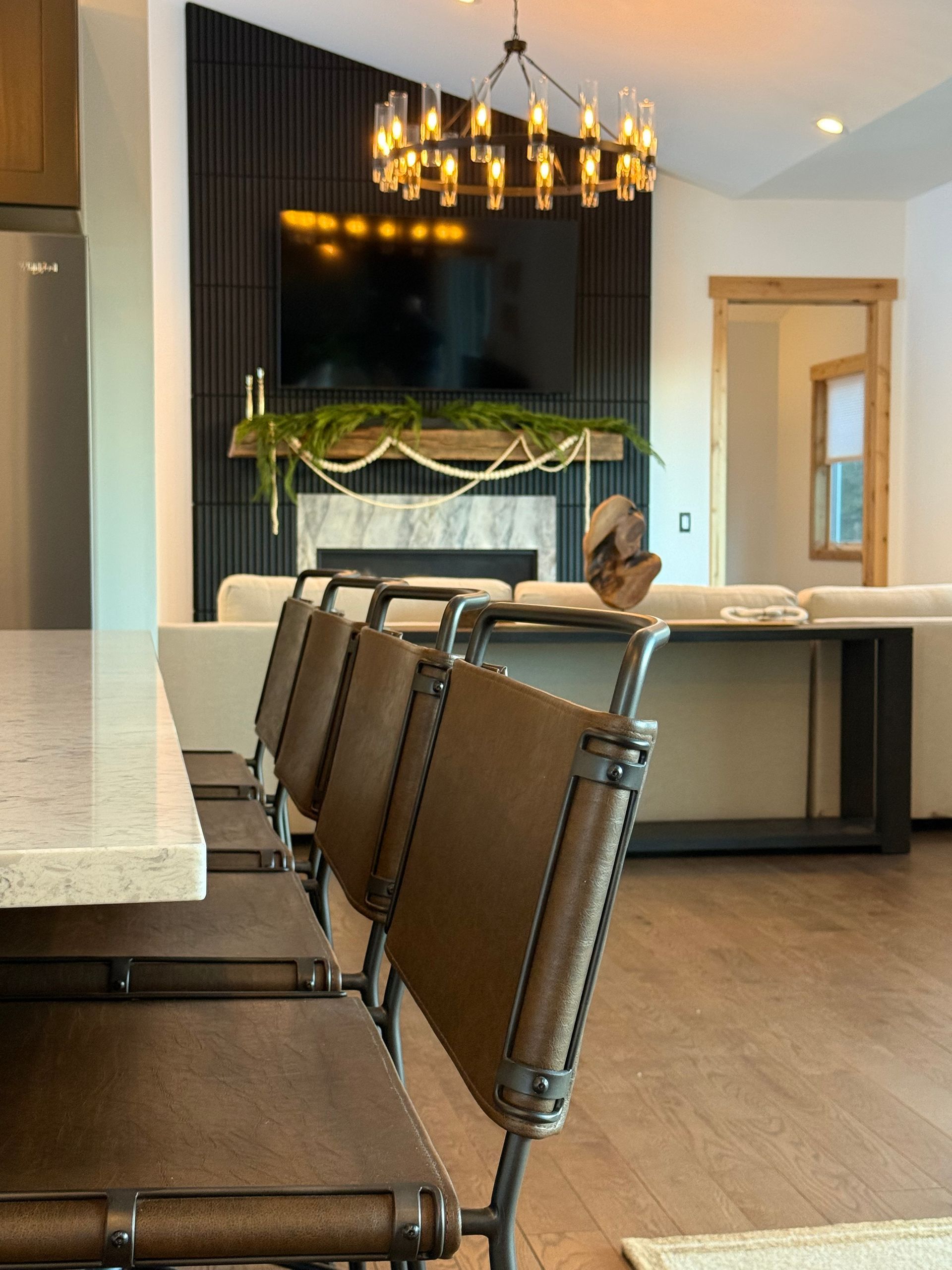 A row of brown chairs at a kitchen counter facing a living room with a fireplace, television, and hanging chandelier.