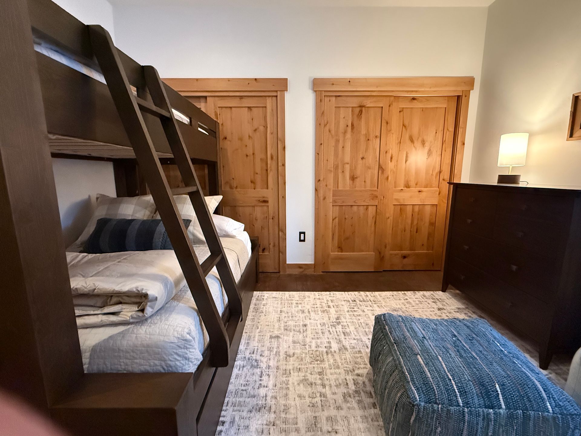 Bunk beds with ladder in a room with wood doors and a blue ottoman.