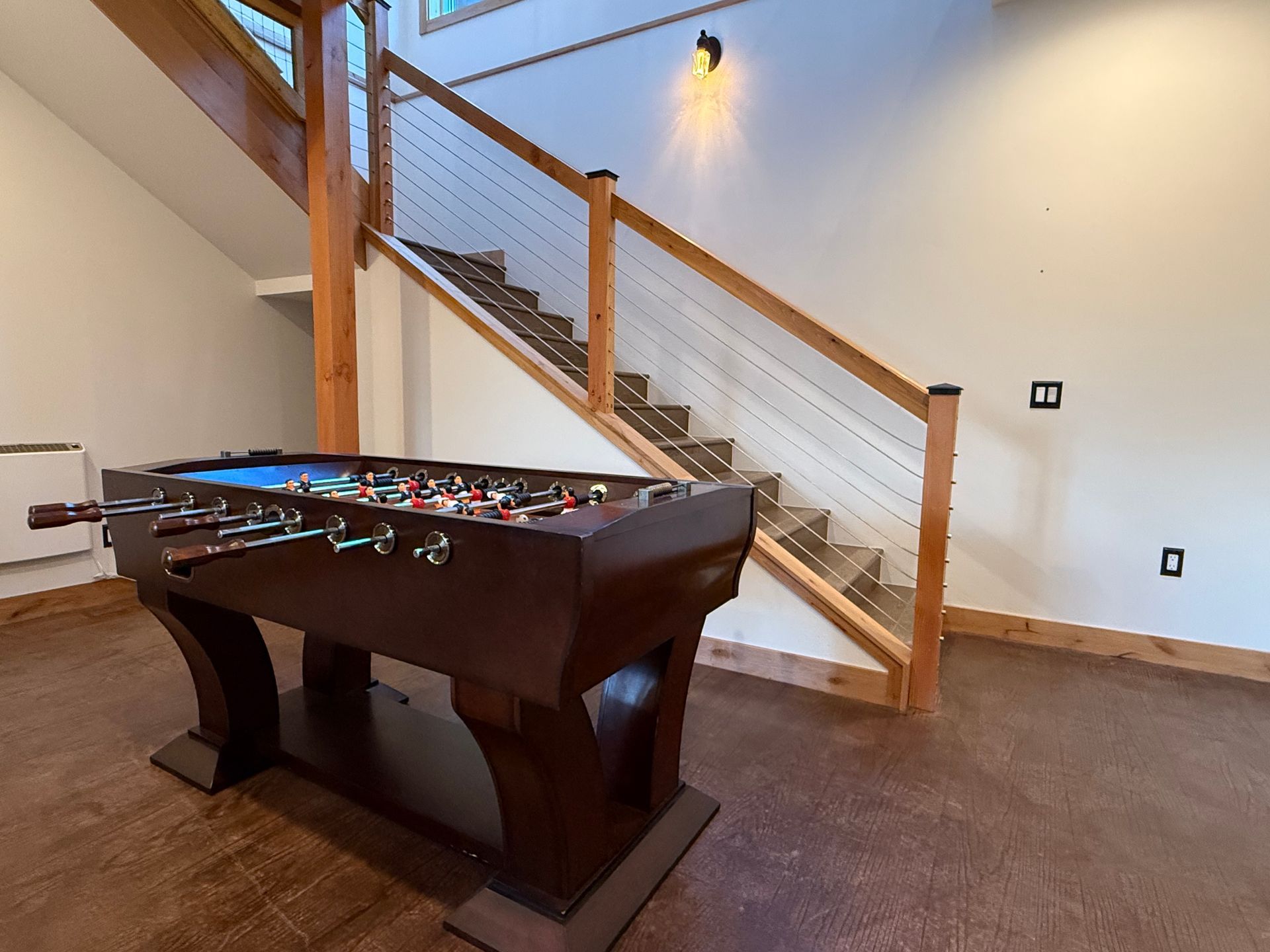 Foosball table under wooden staircase with cable railing, in a room with light brown flooring and white walls.