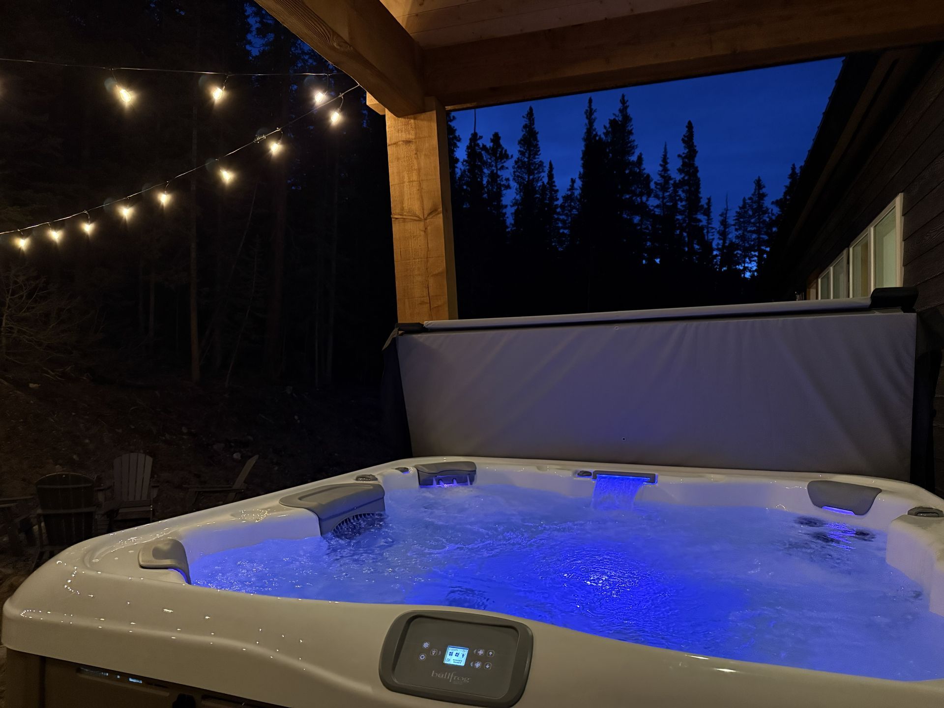 Blue-lit hot tub on a covered patio at dusk, with string lights and trees in the background