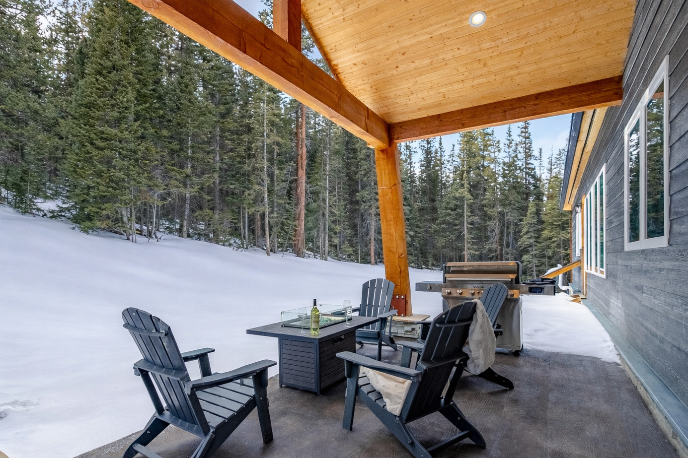 A covered patio featuring two dark Adirondack chairs, a fire pit table, and a grill, overlooking a snowy, pine-forested hill.