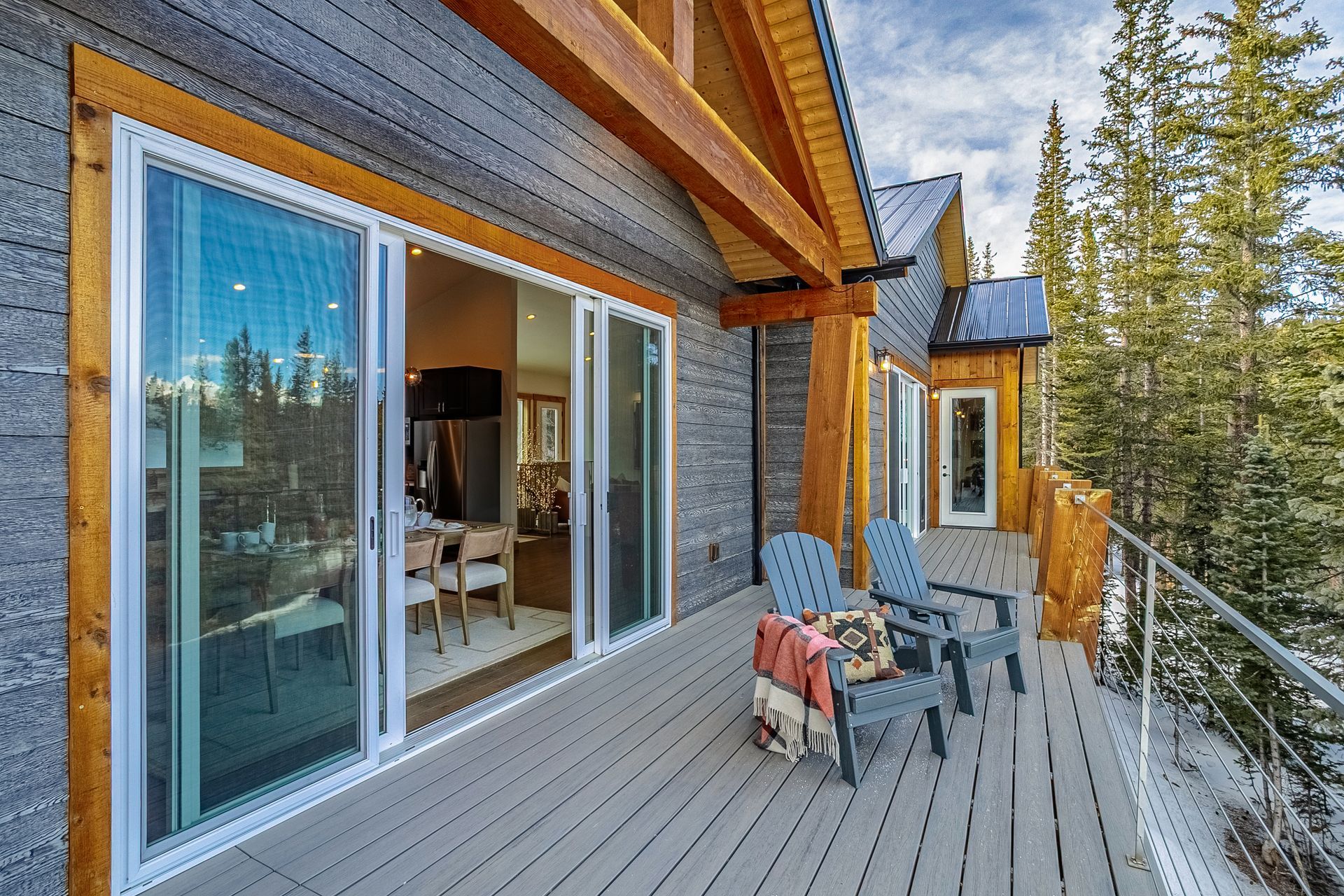 A modern wooden balcony with two blue chairs and sliding glass doors overlooking a snowy, tree-lined landscape.