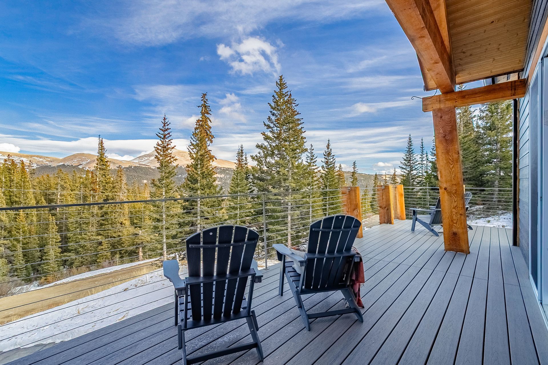 Wooden deck with two chairs overlooking evergreen trees and mountains under a blue sky