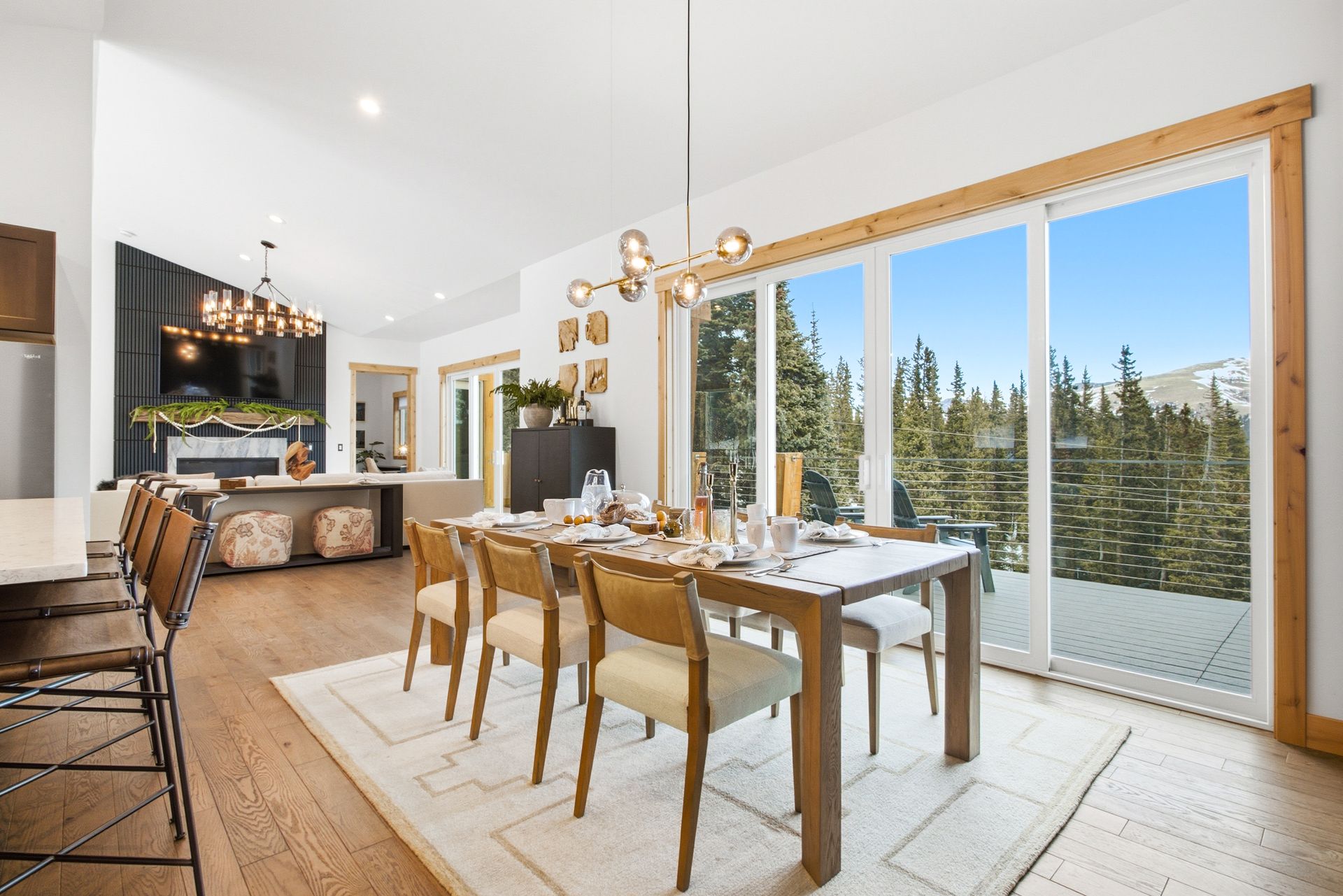 A modern dining room with a wooden table, six chairs, and a chandelier, next to sliding glass doors overlooking a forest.