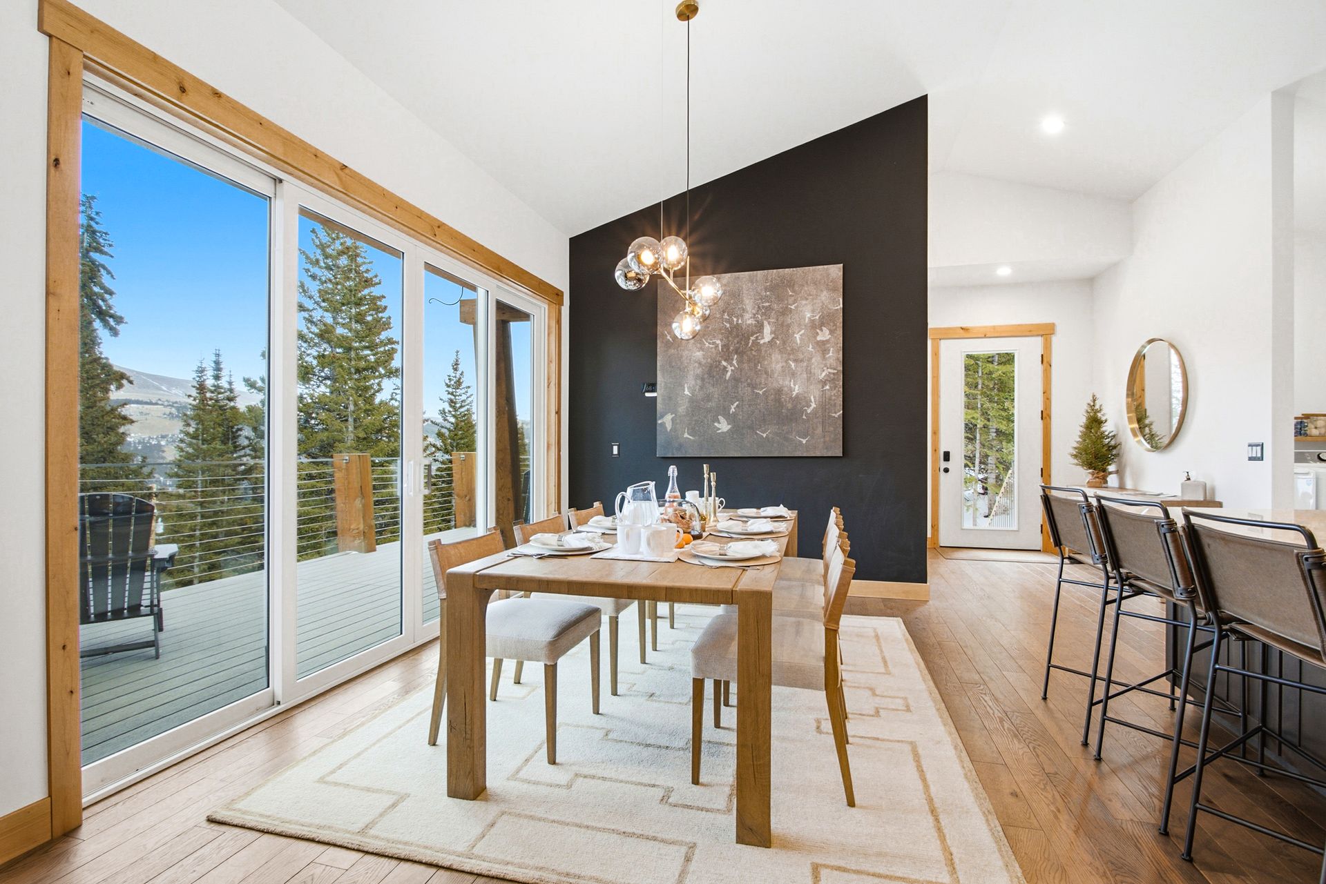 A modern dining room with a wooden table, grey chairs, pendant lights, and large sliding glass doors overlooking trees.