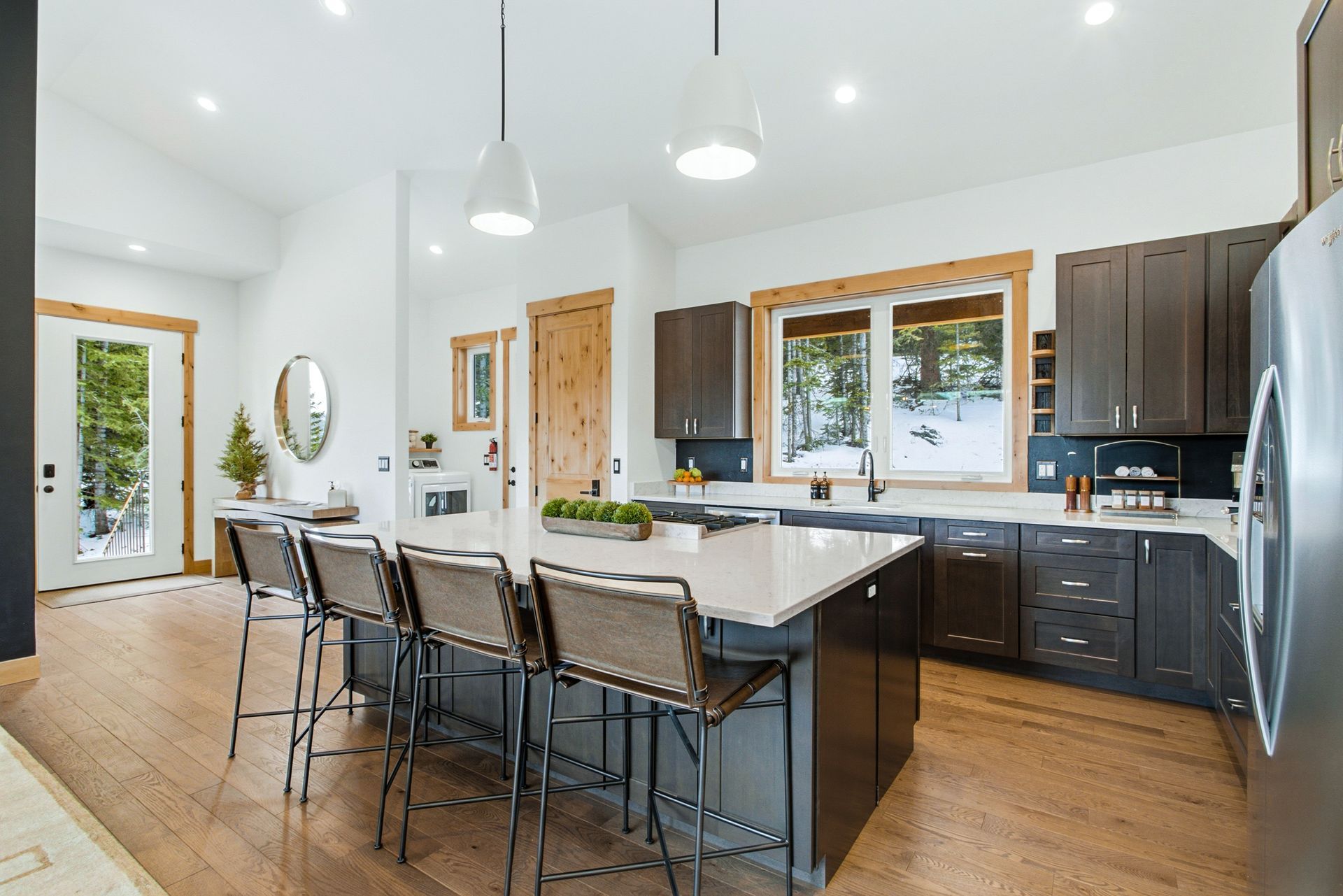 A modern kitchen with a large island, three stools, dark brown cabinets, white countertops, and wood floors.