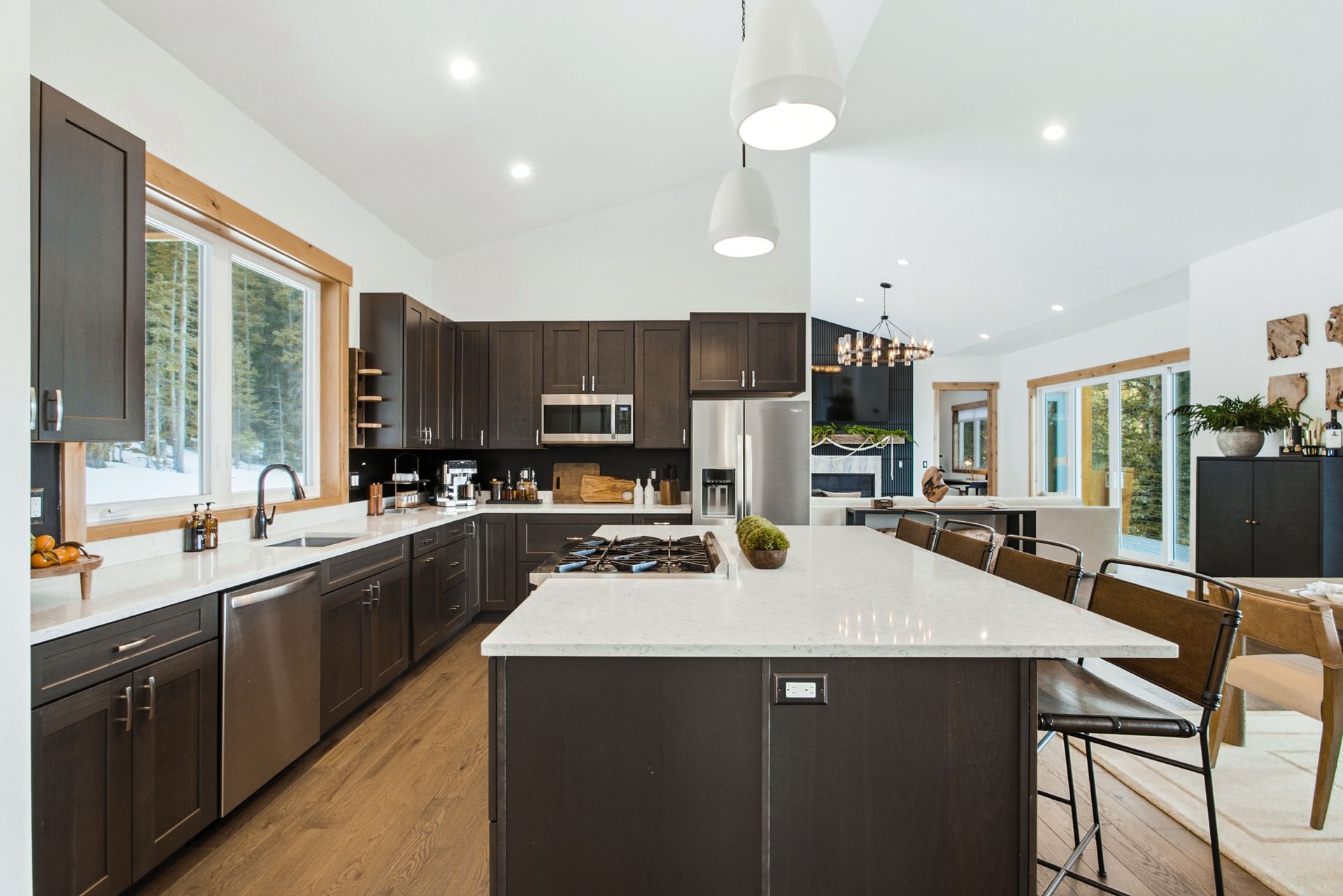 Modern kitchen with dark wood cabinets, a white island, stainless steel appliances, and large windows looking outside.