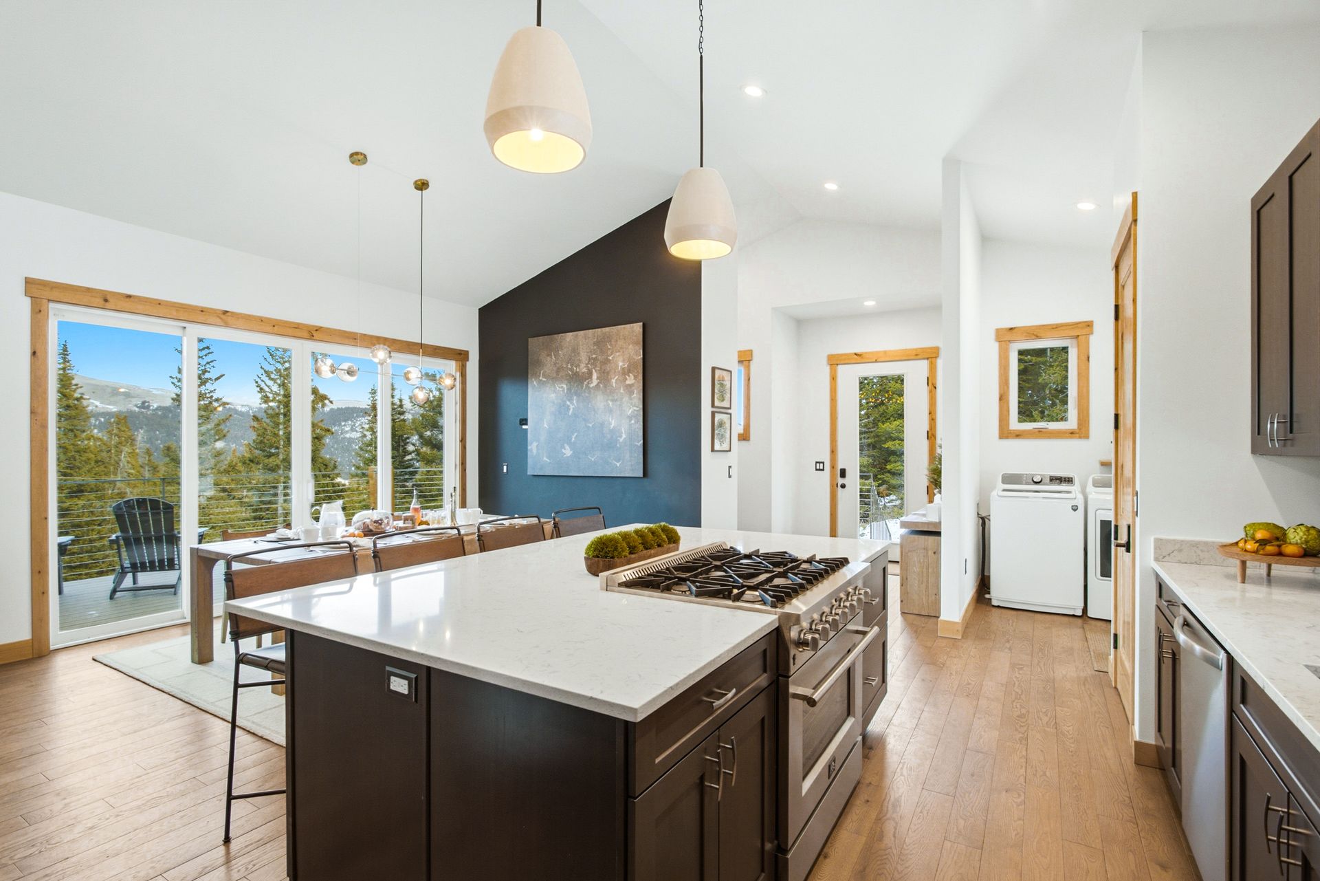 A modern kitchen with a large white island, dark cabinets, and a dining area overlooking a snowy mountain landscape.