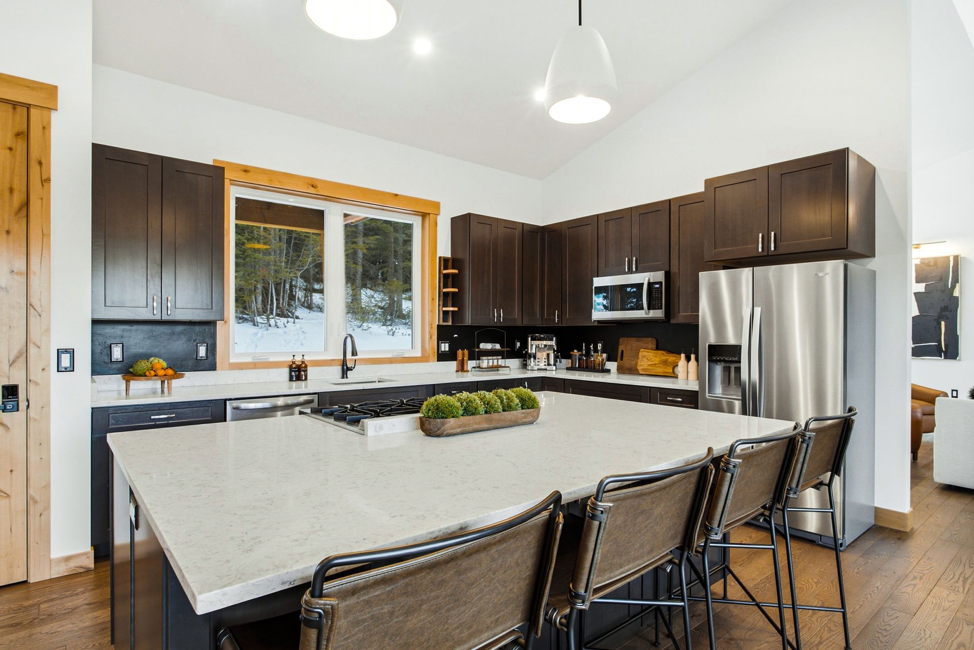 A bright, modern kitchen featuring dark wood cabinets, a large white island with seating, and a window with snowy views.