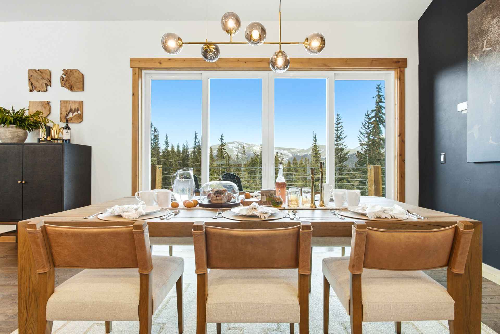 A dining room featuring a wooden table, three upholstered chairs, a modern chandelier, and a large window facing mountains.