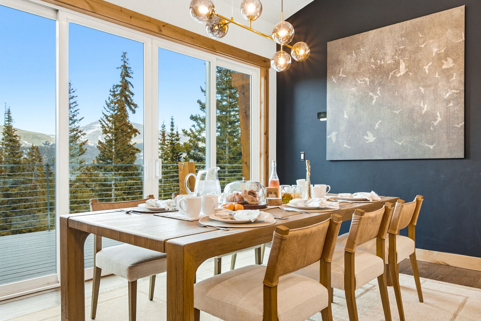 A dining room with a wooden table and chairs, set against a dark blue wall with art, overlooking a mountain forest.