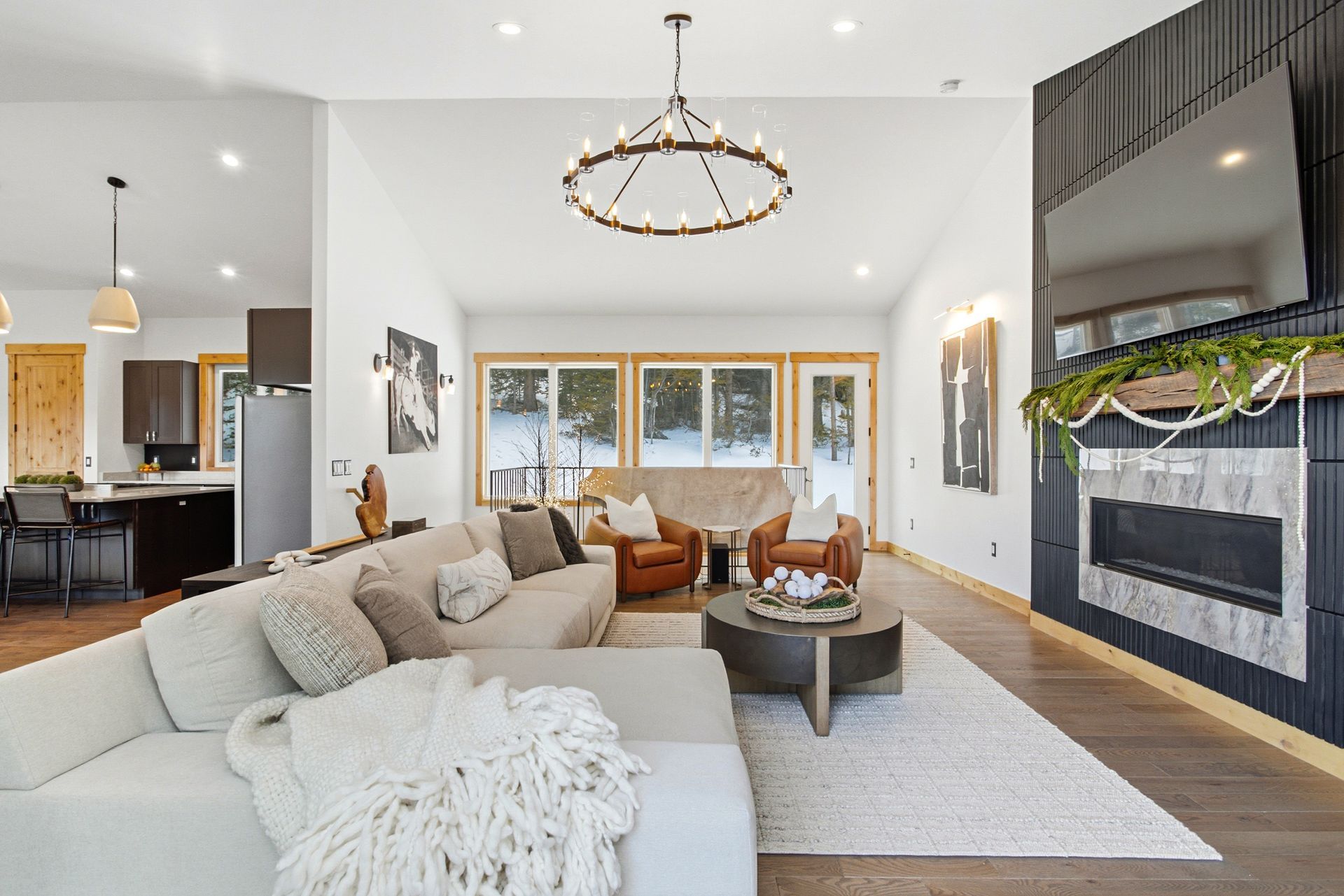 A modern living room with a gray sectional, two leather chairs, a coffee table, fireplace, and open-plan kitchen.