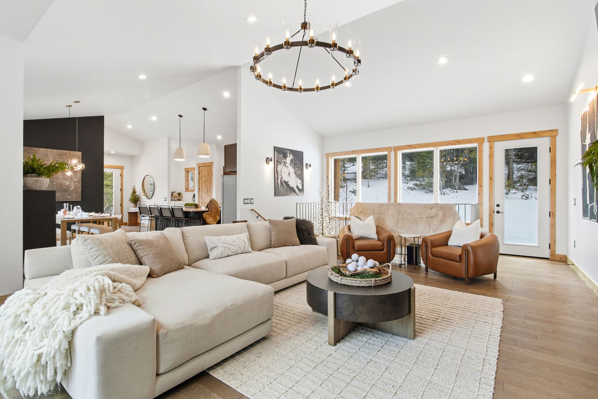 A spacious, modern living room with white walls, a large beige sectional sofa, a round coffee table, and two leather chairs.