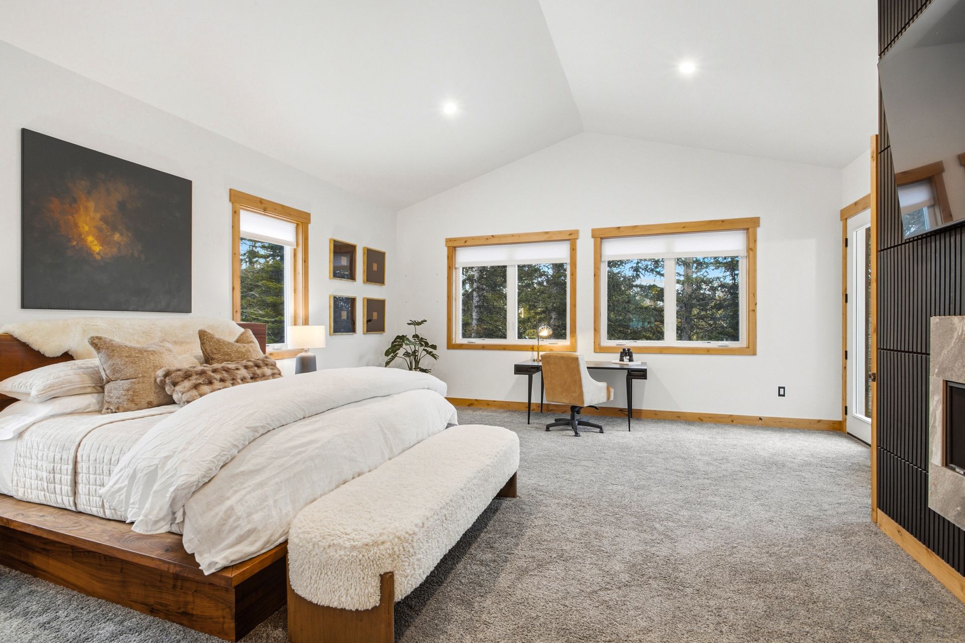 A bedroom with a king bed, cream bedding, a textured bench, dark wood accents, and a desk under windows in a bright room.