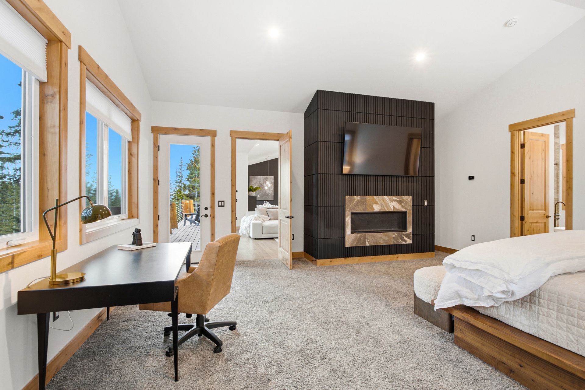 A modern bedroom with a desk, office chair, large fireplace with a mounted television, and a bed with white bedding.