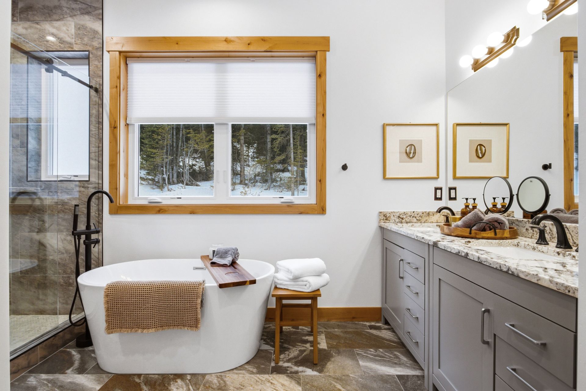 Modern bathroom with a freestanding tub, a walk-in stone shower, a gray double vanity, and a large window facing trees.