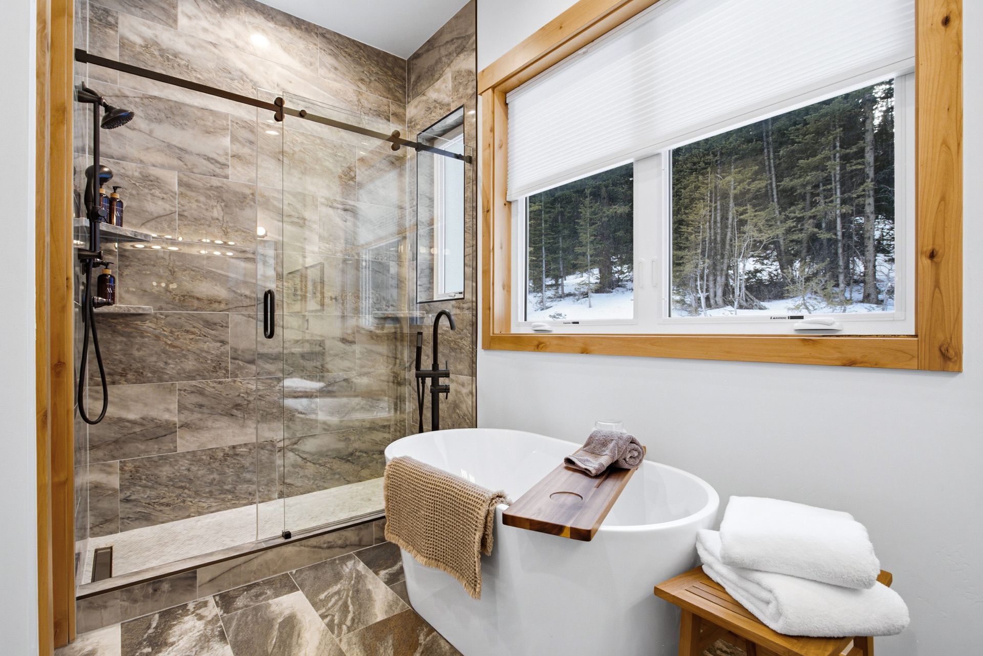 A bright modern bathroom featuring a white soaking tub next to a large window and a stone-tiled walk-in glass shower.