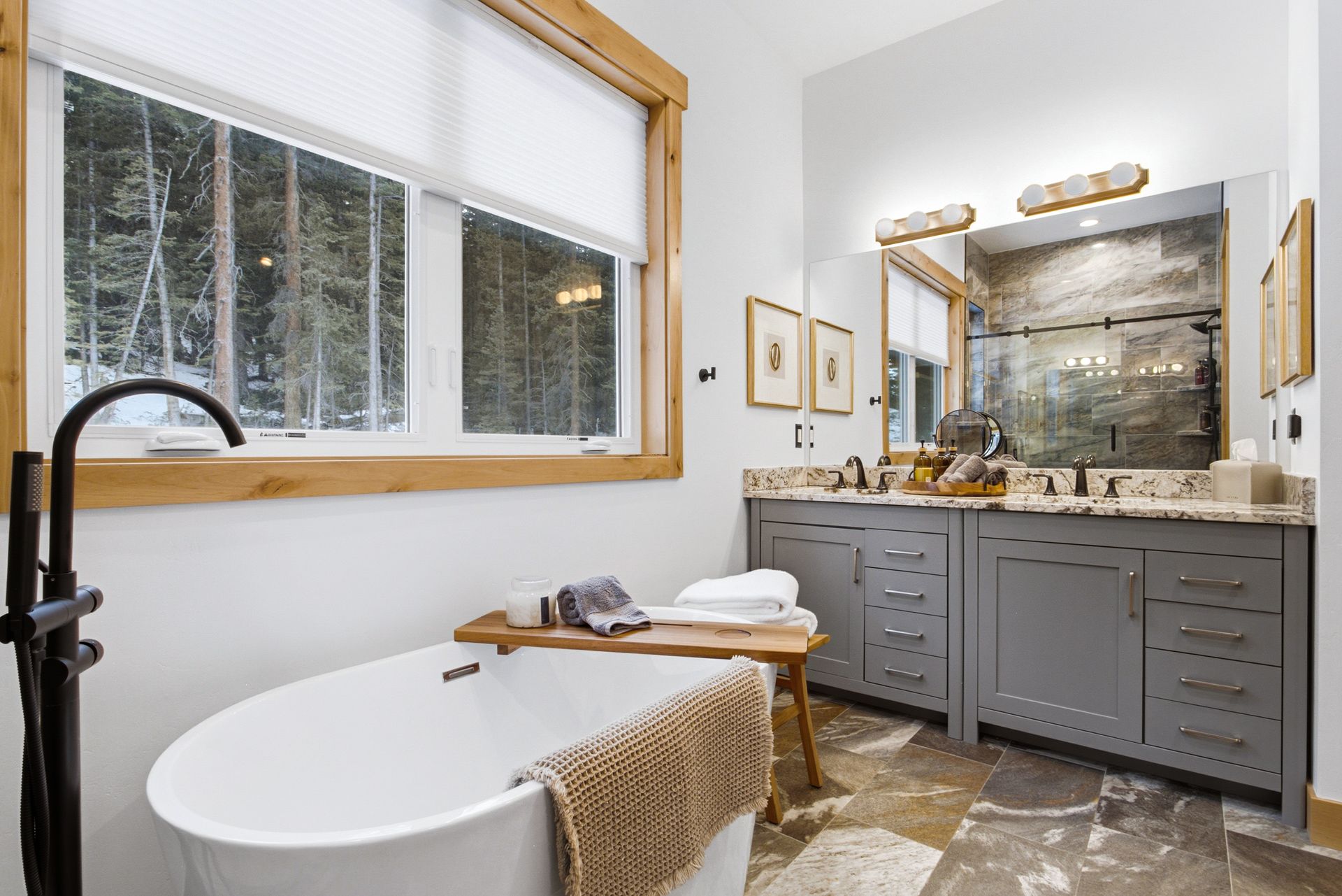 Modern bathroom with a freestanding white tub, grey vanity, stone flooring, and a large window overlooking a forest.