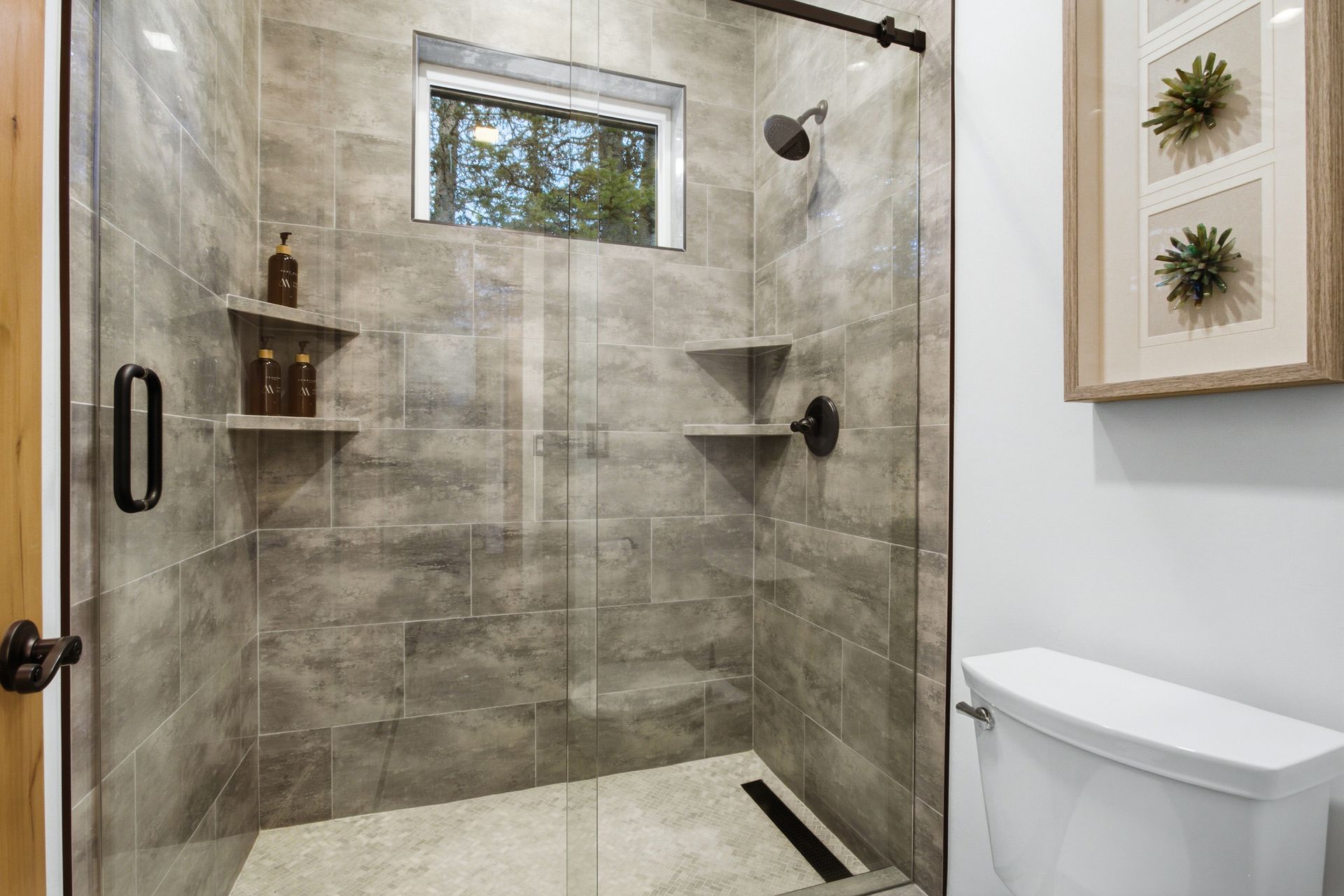 A modern walk-in shower with grey stone-look tile, glass door, built-in shelves, and a toilet in a white bathroom.