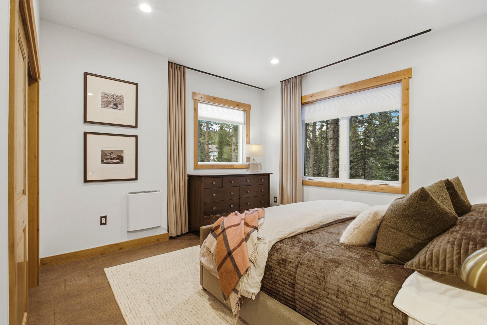 A cozy, well-lit bedroom featuring a bed with brown bedding, wooden accents, two framed prints, and a windowed dresser.