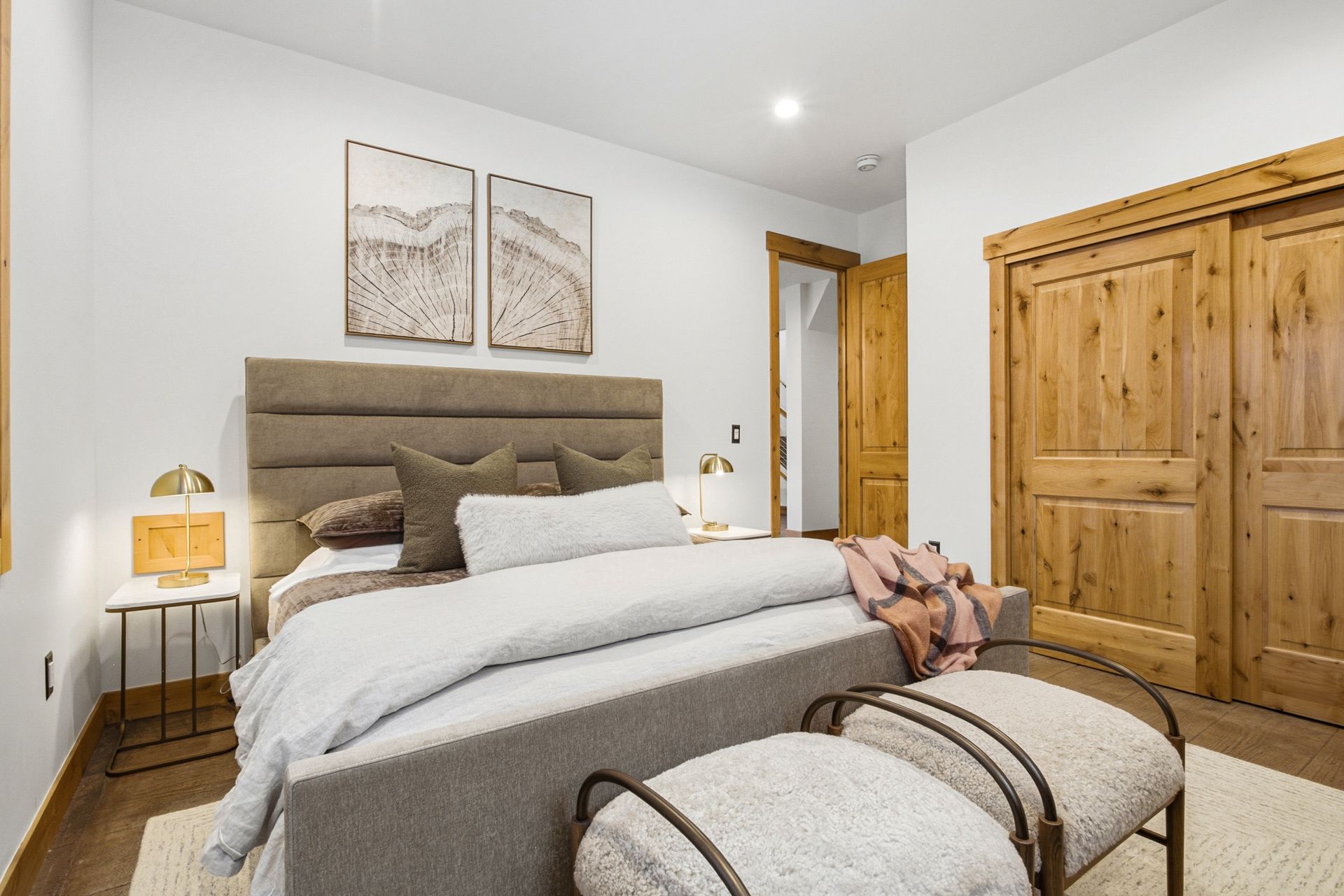 A modern bedroom features a gray upholstered bed, light bedding, two framed prints, wood sliding doors, and side benches.
