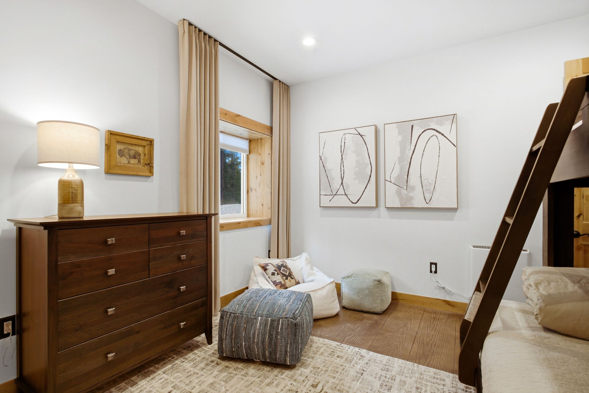 A bedroom with a wooden dresser, a window with beige curtains, a patterned ottoman, a beanbag chair, and bunk bed stairs.