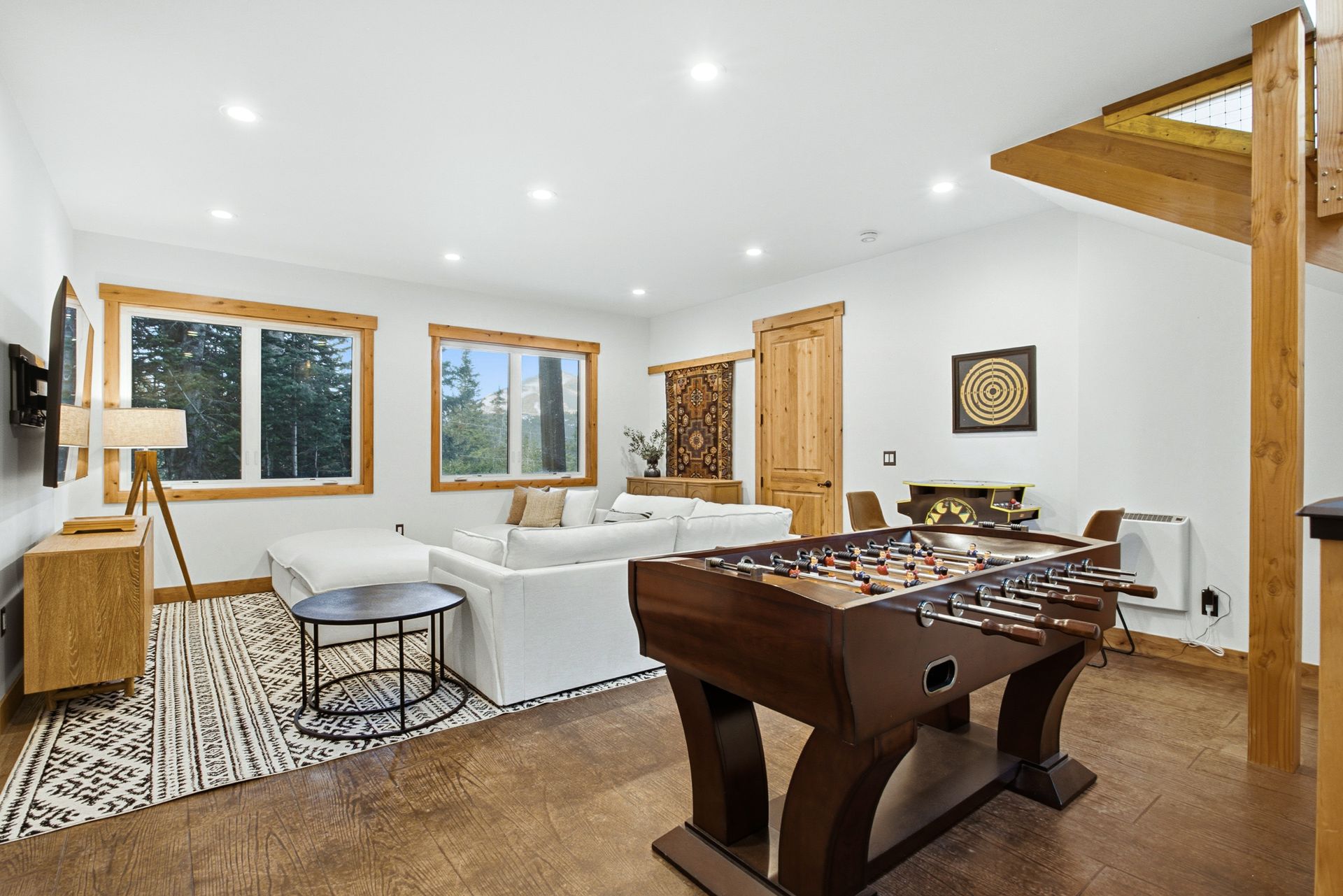 A bright, modern basement lounge with a foosball table, white sofa, patterned rug, and wooden accents.