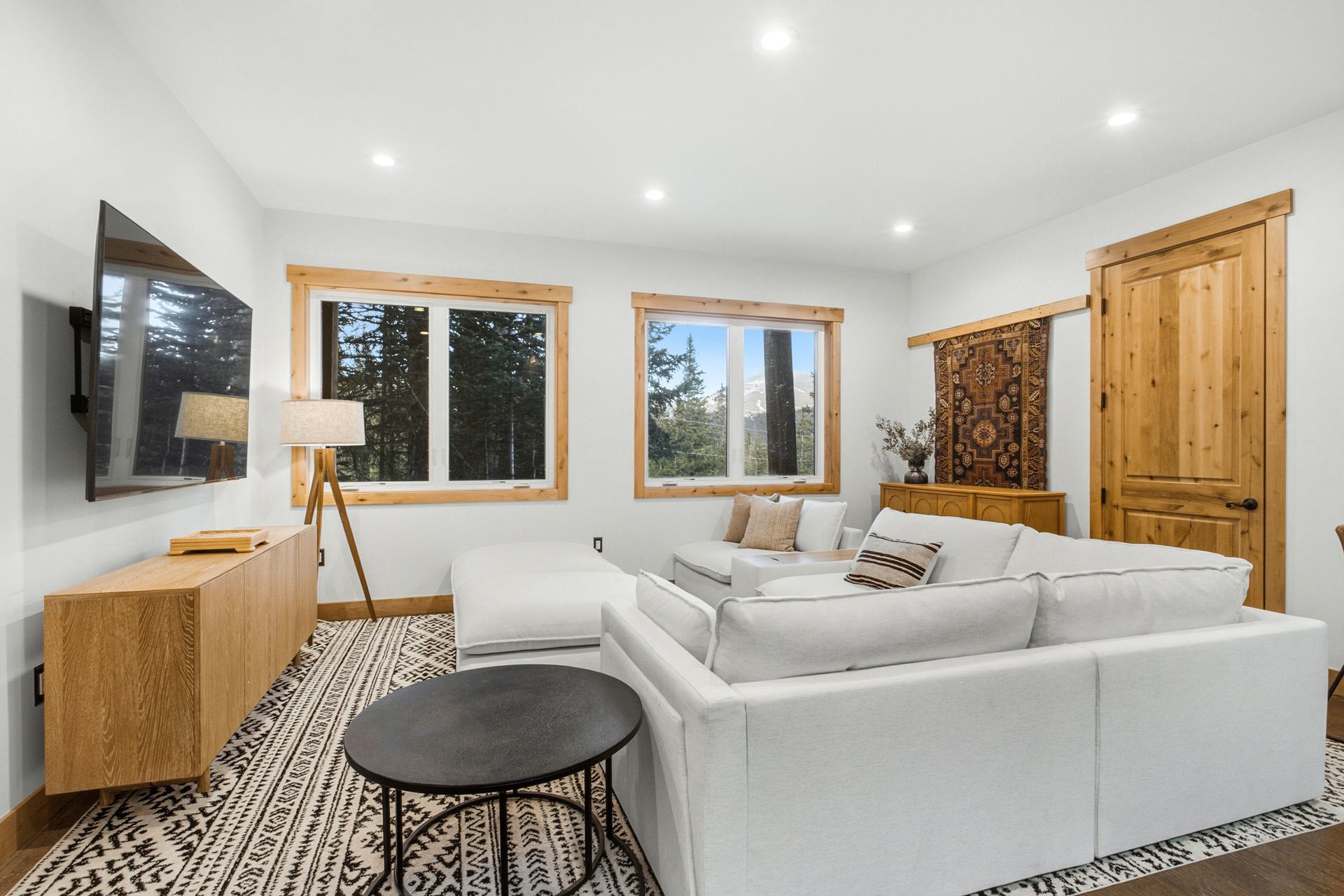 A bright living room with a white sectional sofa, a patterned rug, a wooden media console, and two windows with wood trim.