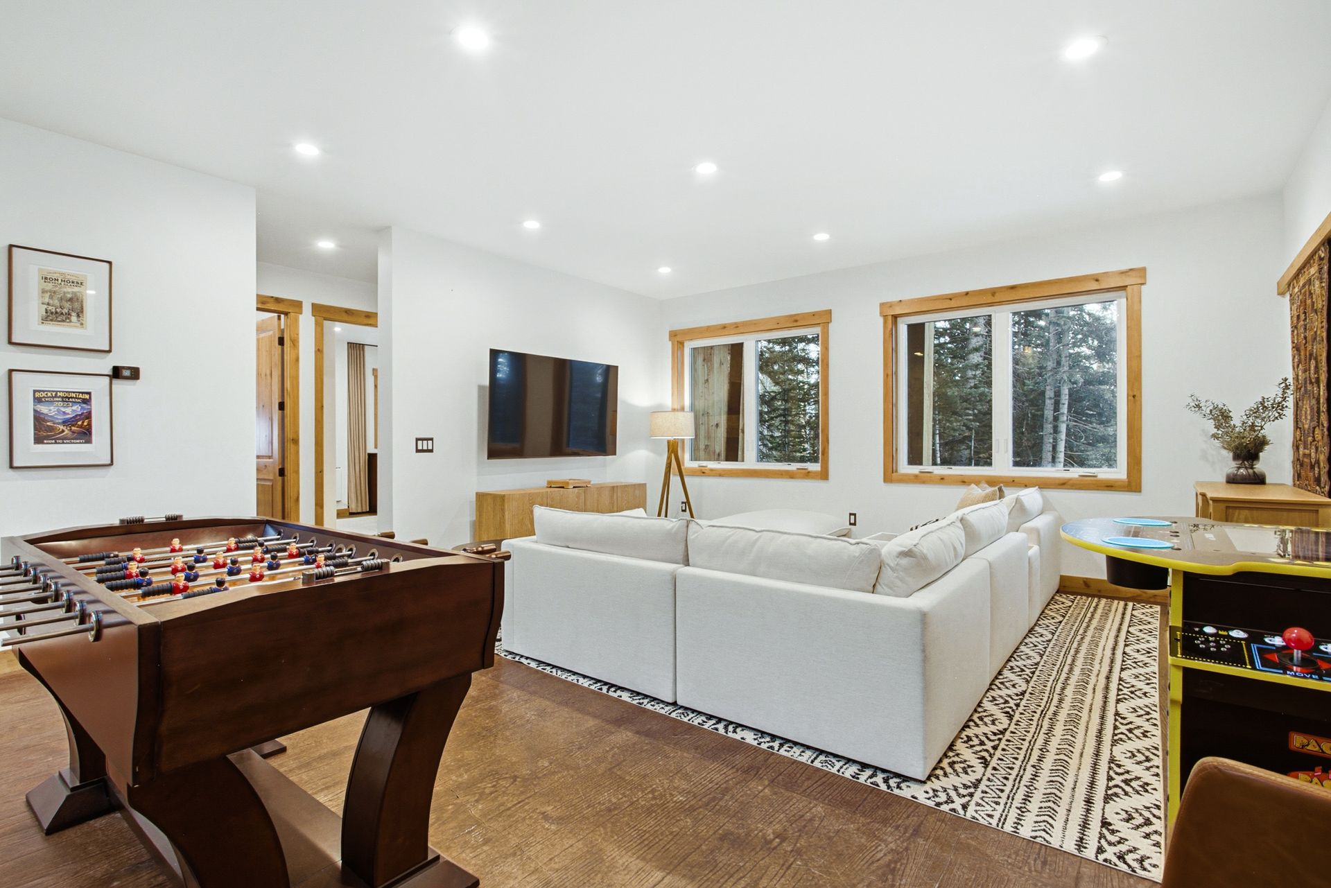 A modern living room with a white sectional sofa, a foosball table in the foreground, and large windows.