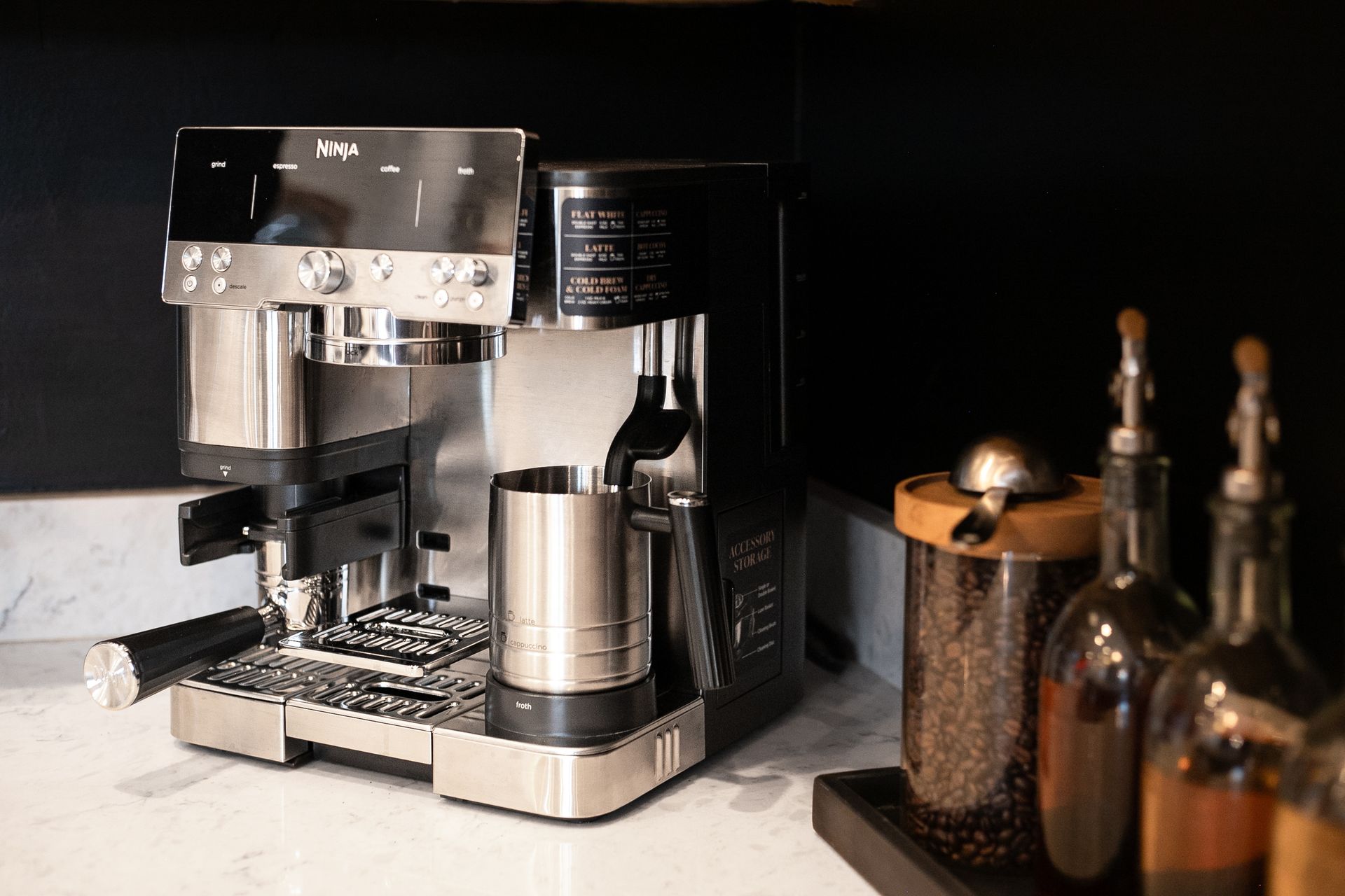 Silver espresso machine on a countertop beside jars and bottles in a dark kitchen setting