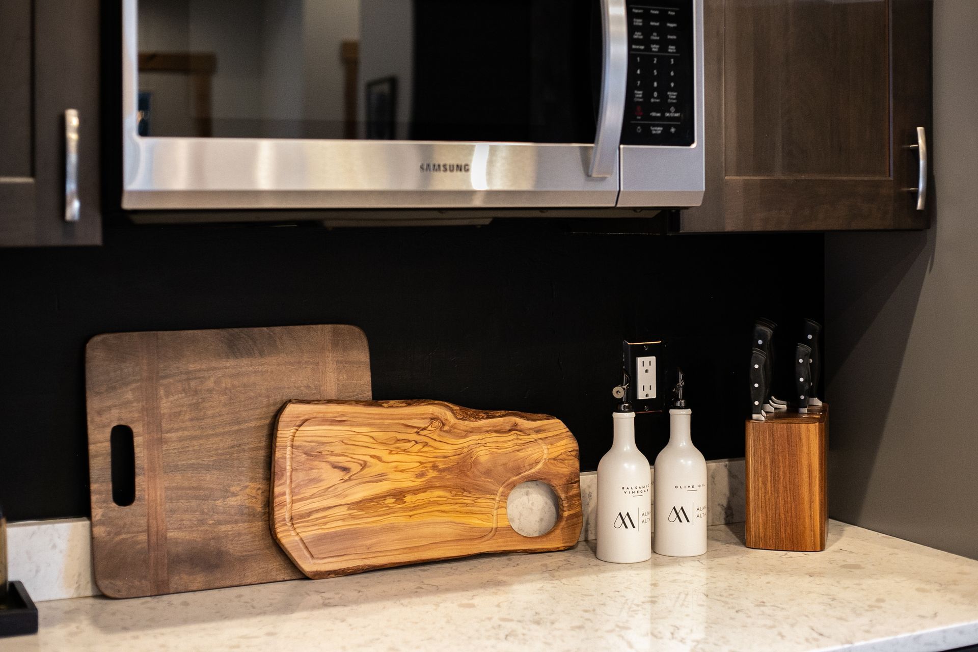 Kitchen counter with two wooden cutting boards, two white oil bottles, and a wooden knife block under a microwave.
