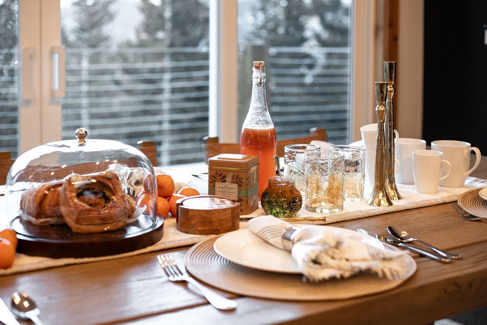 A breakfast table setting with pastries under a glass dome, orange juice, tea, and place settings near a bright window.