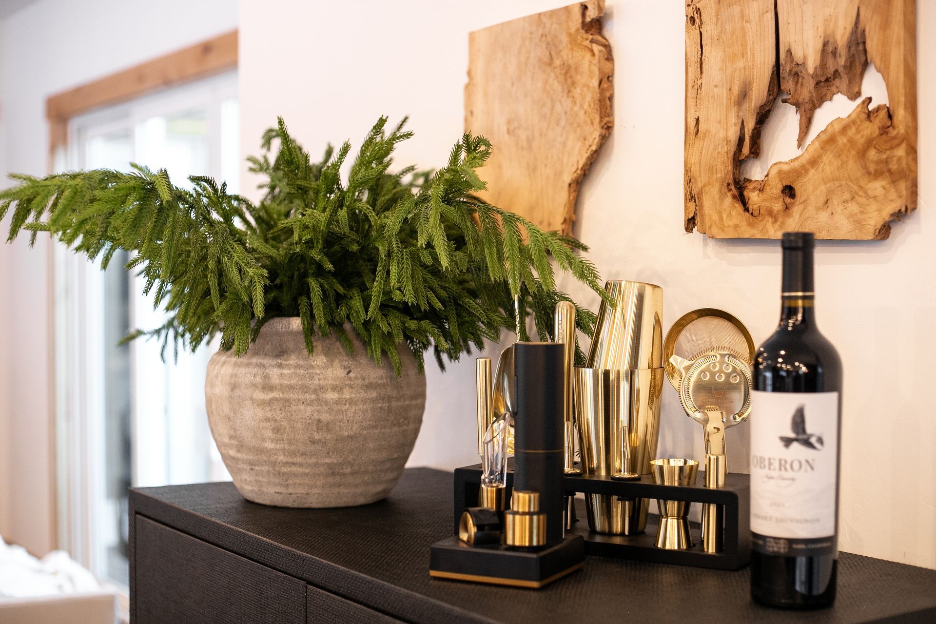 A potted fern, gold bar tools, and a bottle of Heron wine sit on a black table against a wall with wood wall art.