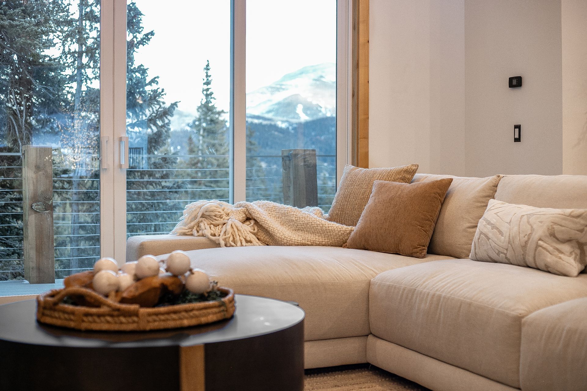 A cozy, neutral-toned sofa with textured pillows faces a large window showing a snowy mountain landscape.