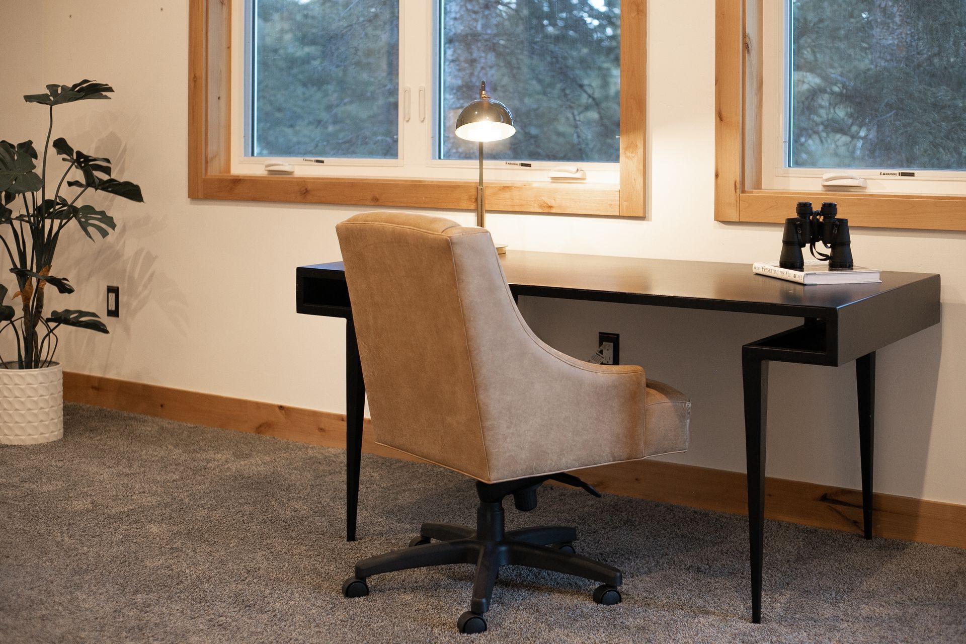 A modern workspace with a tan upholstered chair, a black desk with a lamp and binoculars, against a white wall.