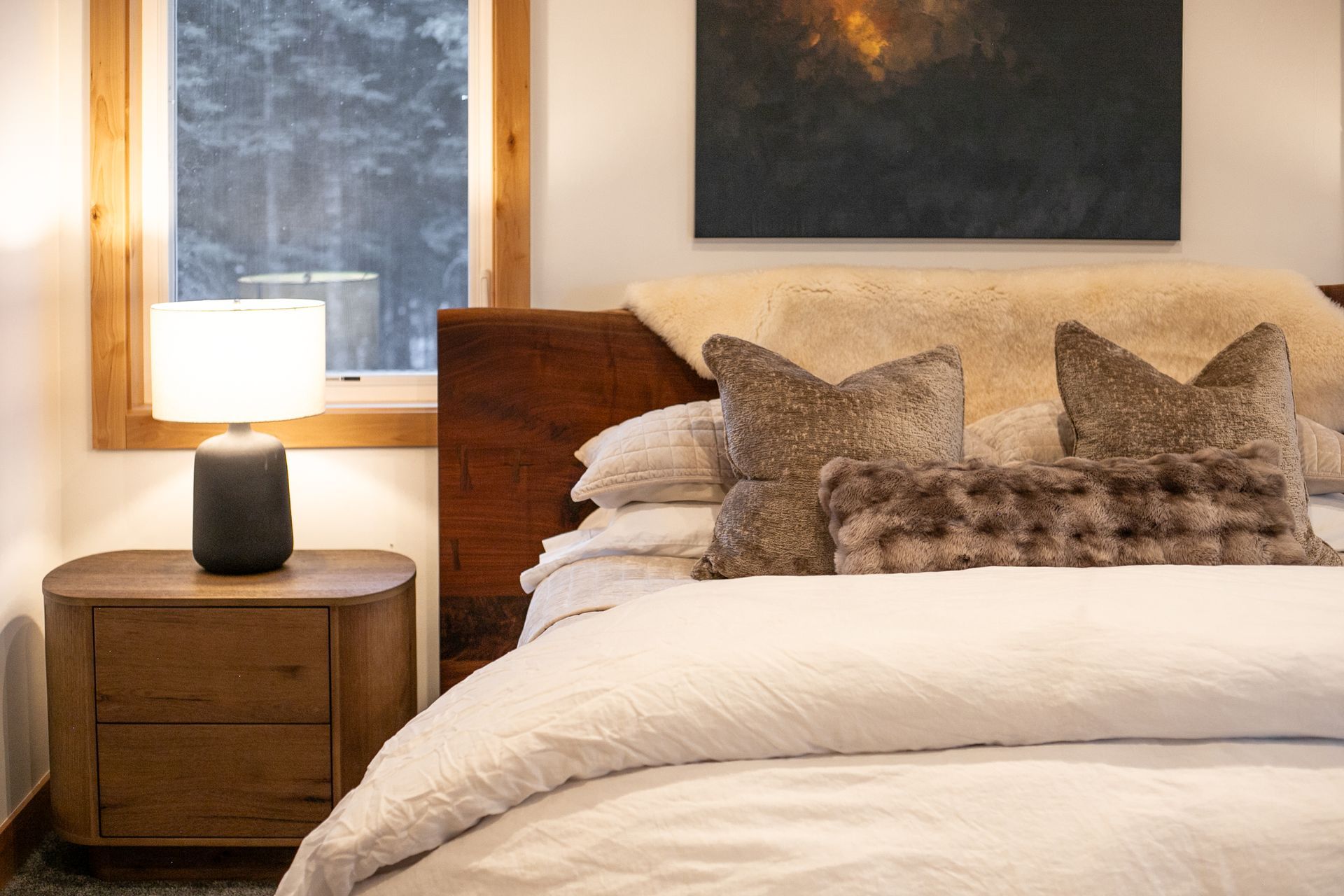 A cozy bedroom with a wooden nightstand, a lit table lamp, and a made bed featuring a white duvet and decorative pillows.