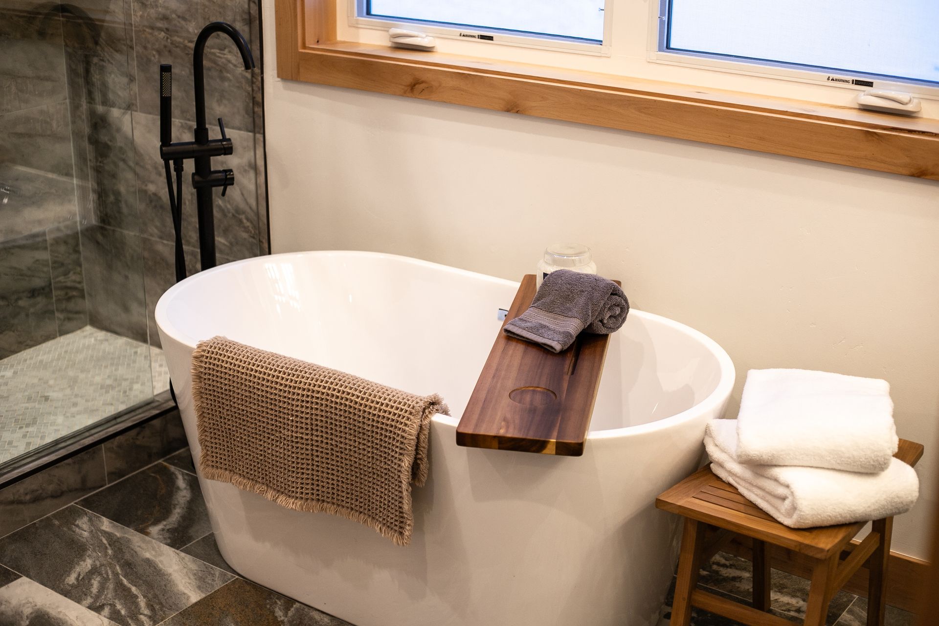 A white freestanding bathtub sits in a bathroom with dark tiled flooring, a wooden tray, and a stool with white towels.