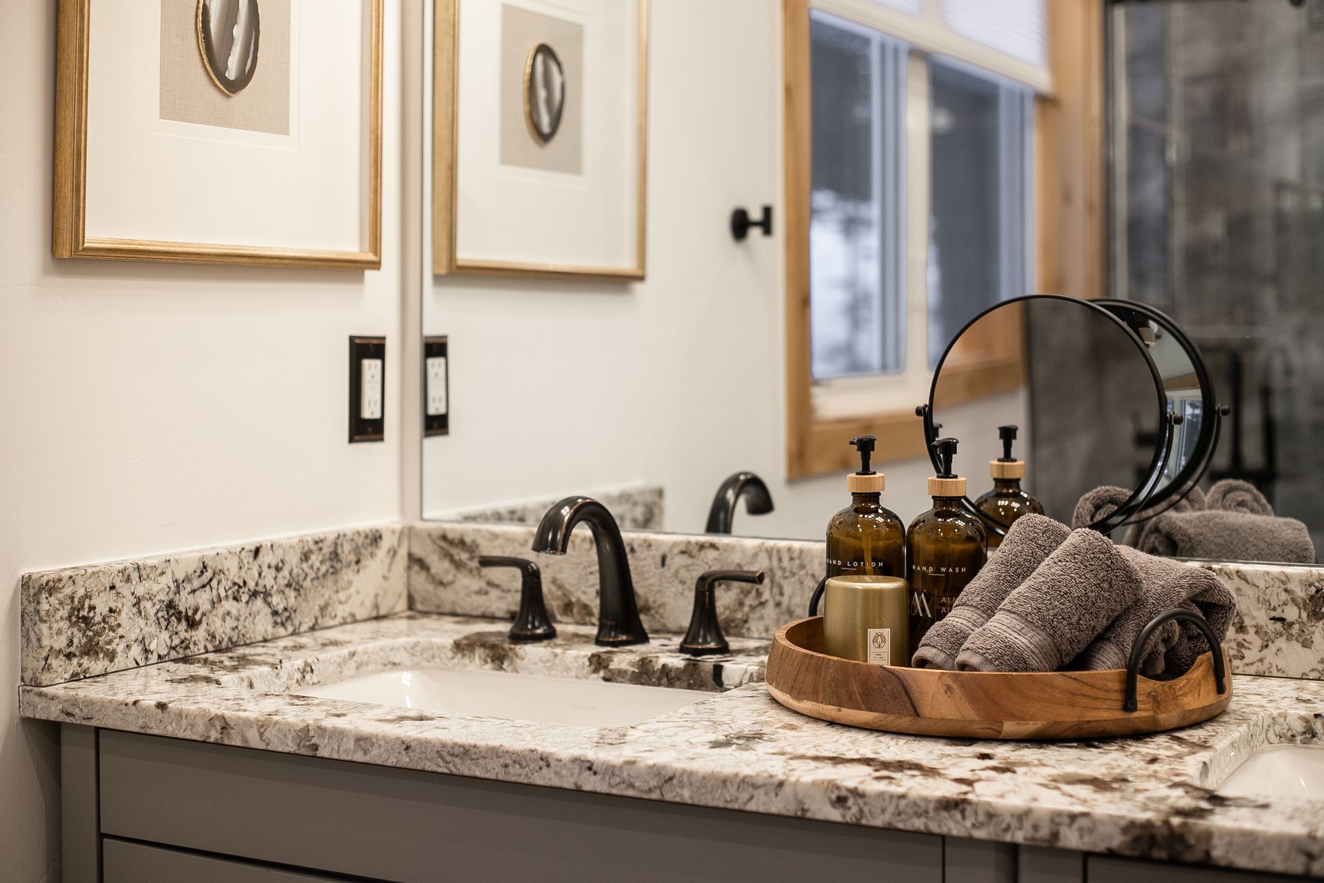 A granite bathroom countertop with a dark faucet, a round vanity mirror, and a tray holding toiletries and grey towels.