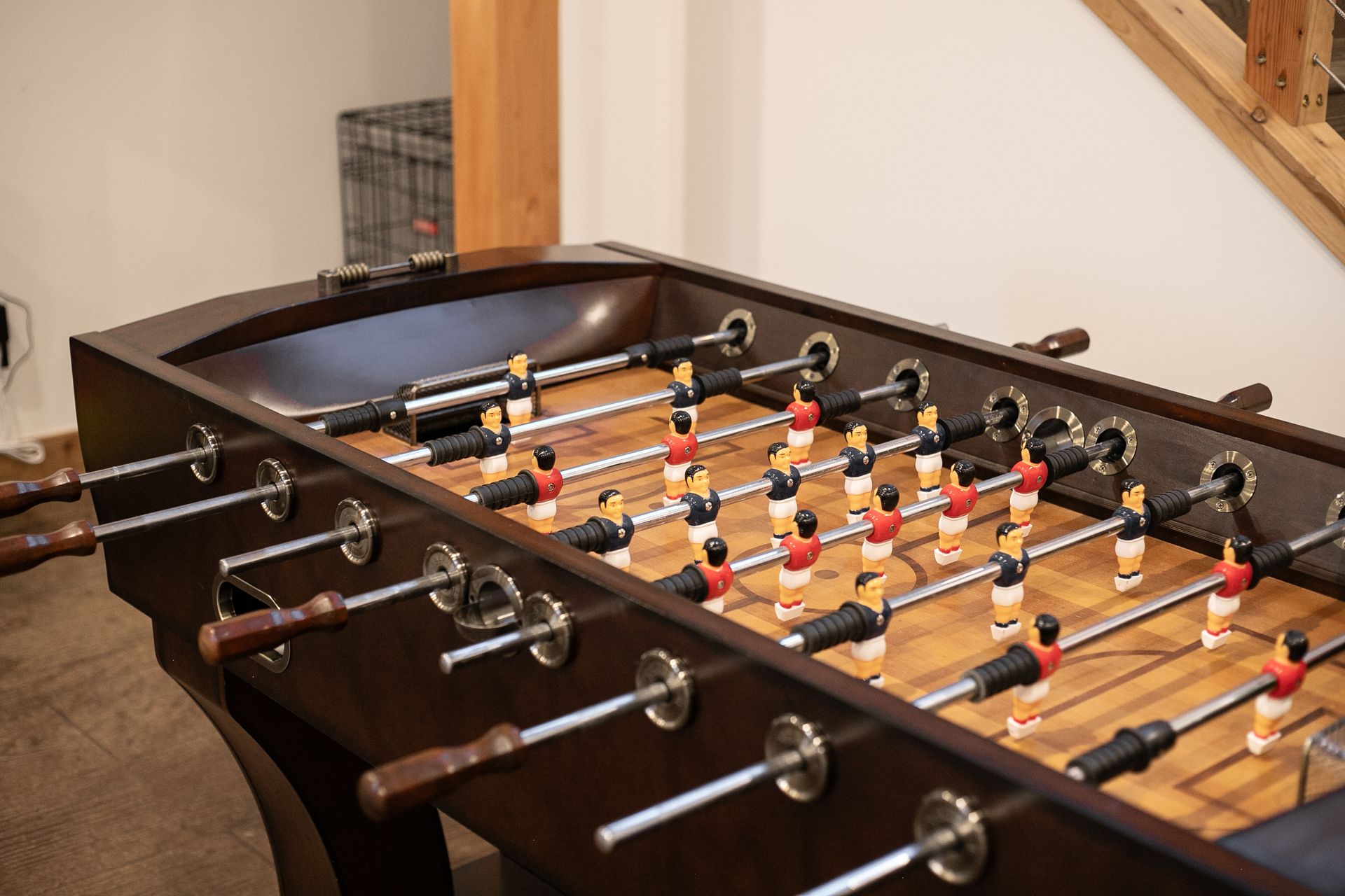 A wooden foosball table with red and black player figurines set on metal rods in a room with light-colored walls.