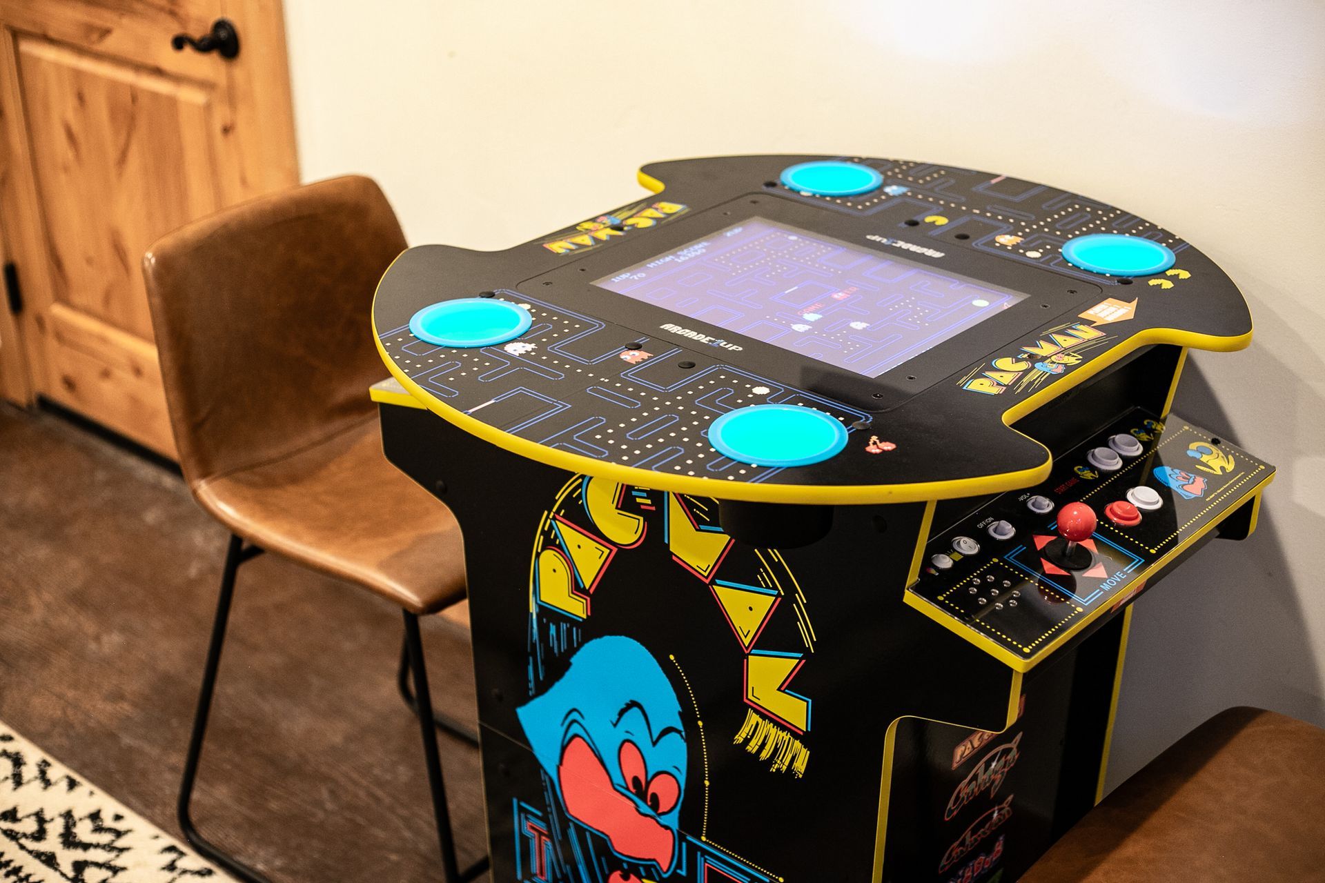 A Pac-Man arcade cocktail table with blue illuminated buttons, situated in a room next to a brown chair.