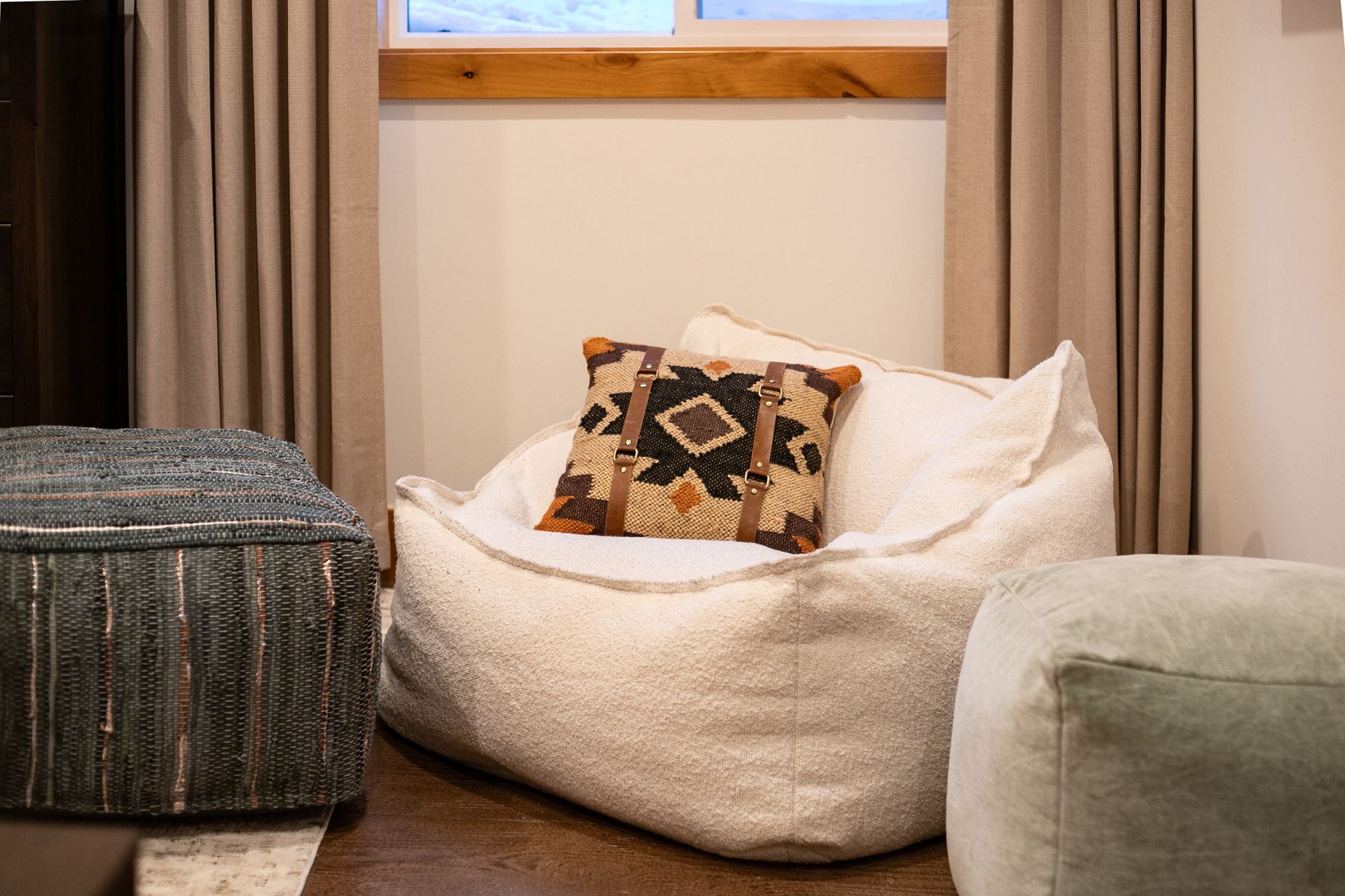 A fluffy white beanbag chair with a patterned throw pillow sits between two fabric ottomans in front of beige curtains.
