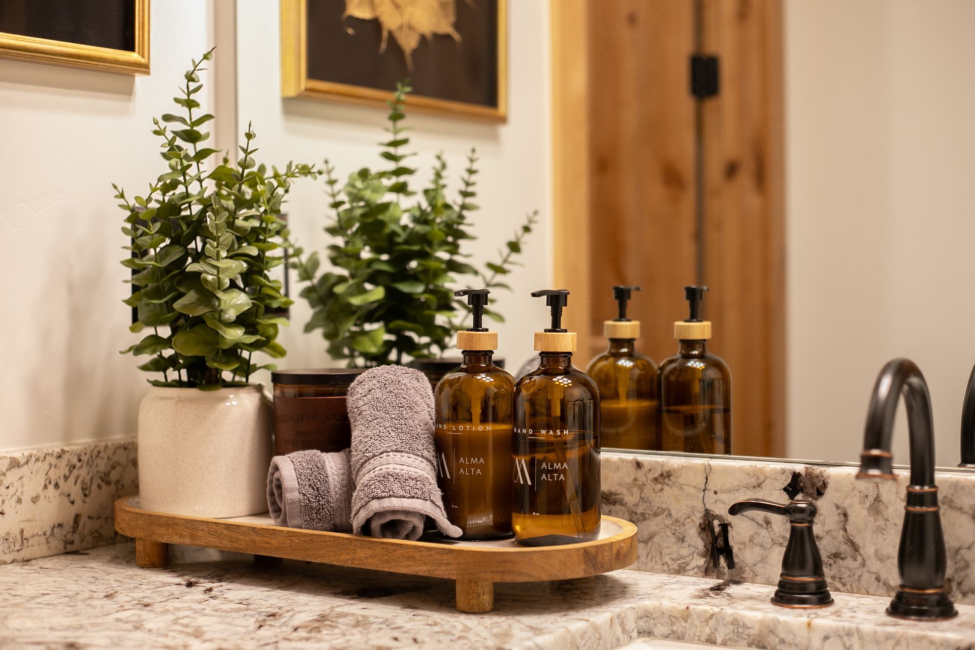 Amber soap bottles, rolled towels, and two potted plants rest on a wooden tray atop a marble bathroom vanity.