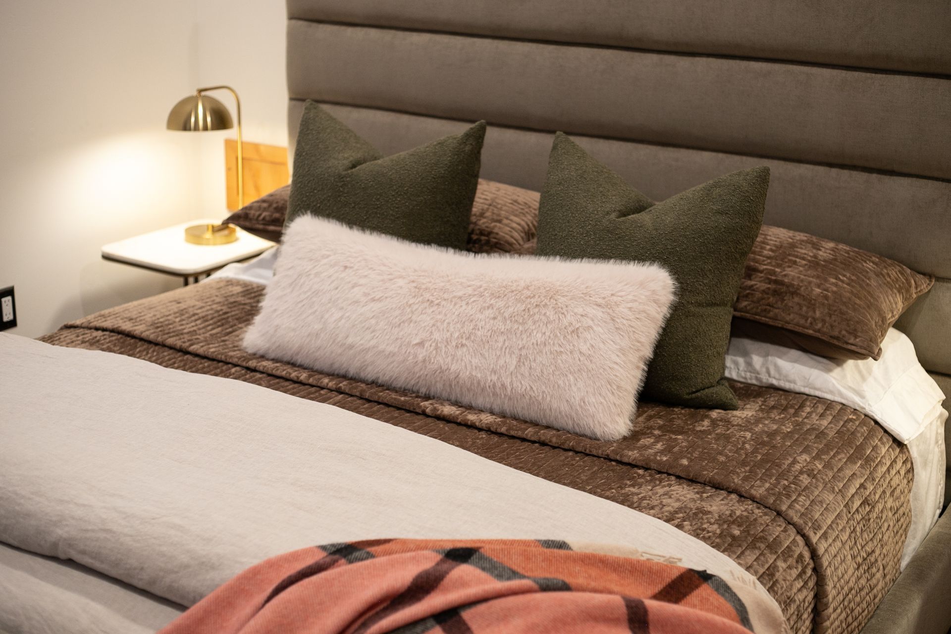 A bed with a tufted headboard, two dark olive pillows, a fuzzy cream lumbar pillow, and a patterned peach-toned blanket.