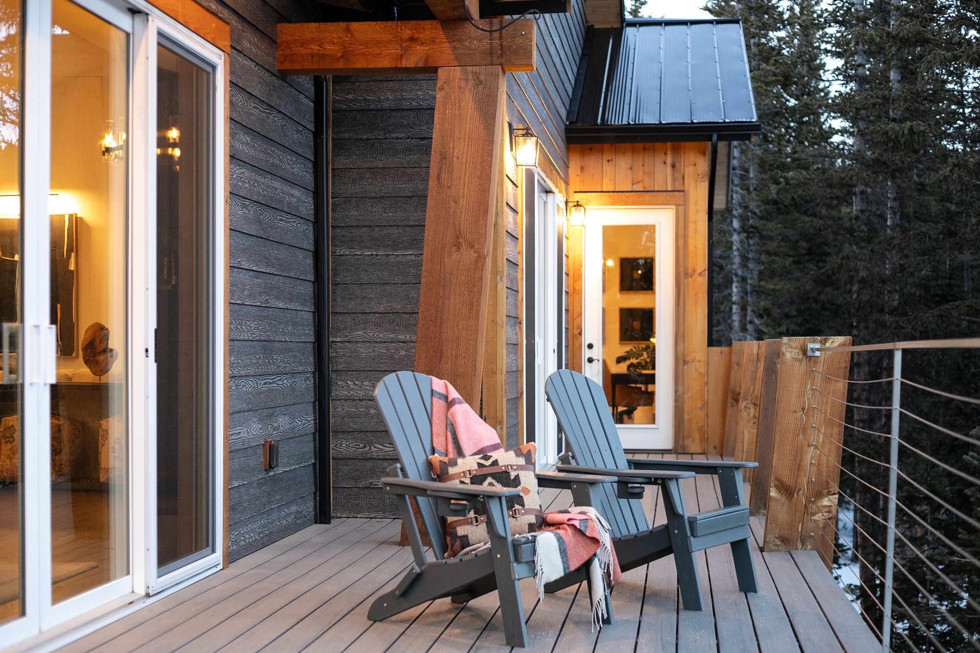 Two Adirondack chairs with blankets sit on a wooden deck outside a modern house with wood siding, overlooking a forest.