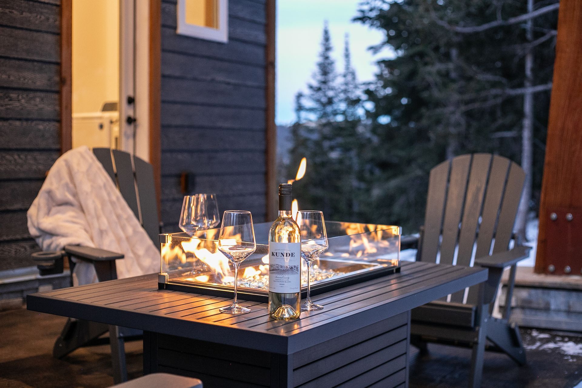 Two Adirondack chairs sit on a deck flanking a fire table with a wine bottle and two glasses, overlooking a snowy forest.