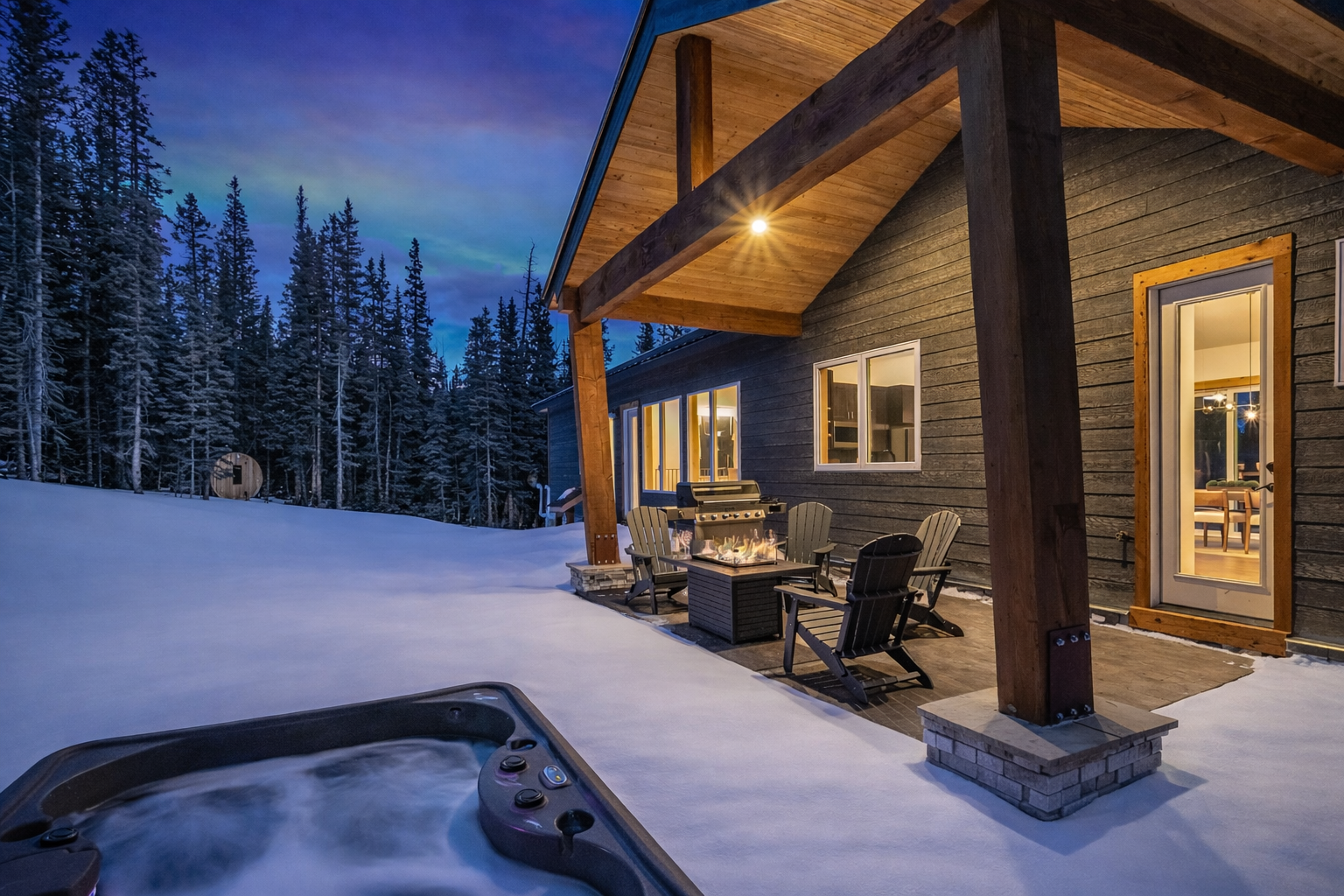 A snowy patio at dusk with a hot tub in the foreground, a fire pit, lounge chairs, and a rustic home against a forest.
